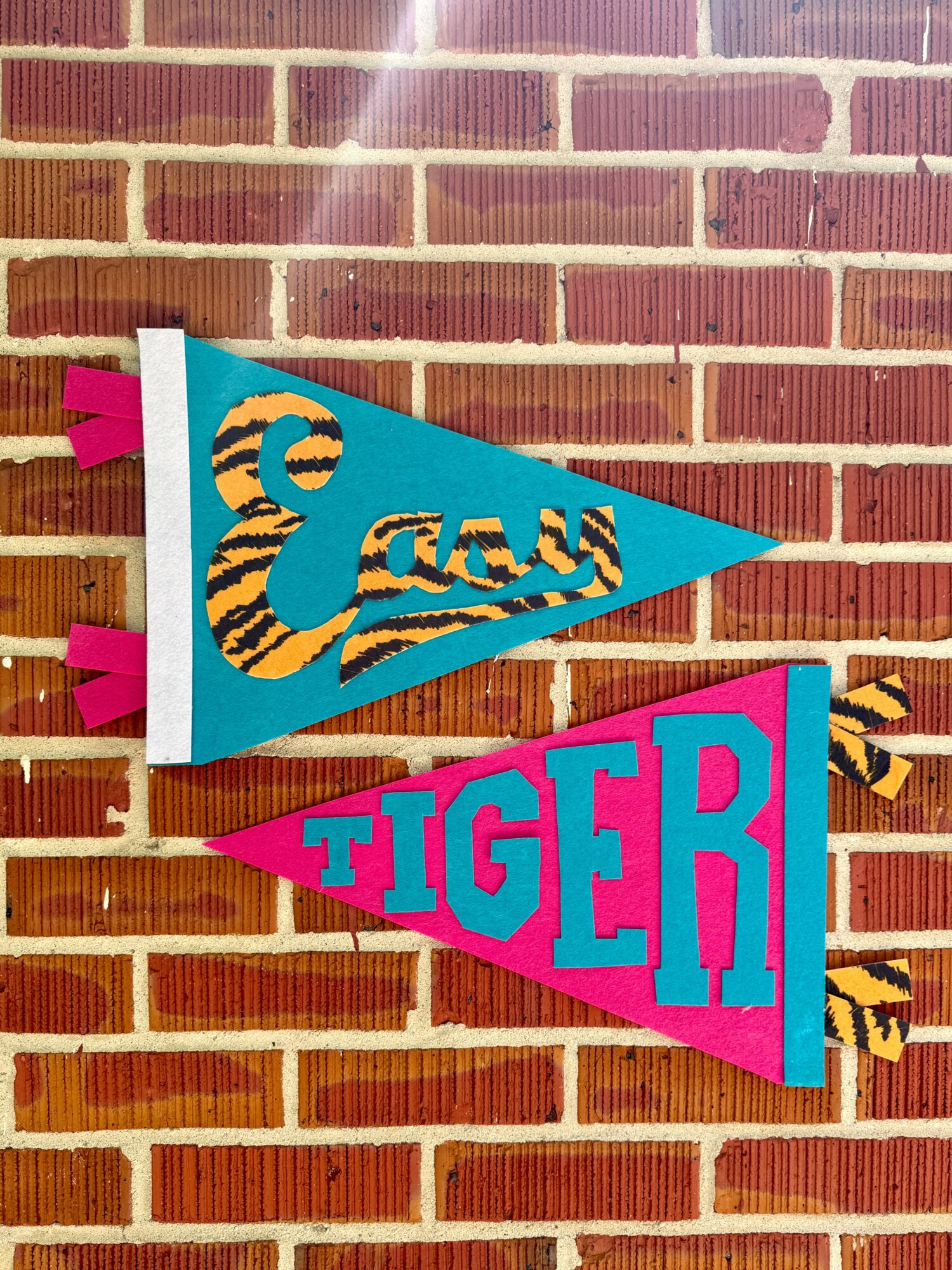 Two colorful pennant banners hanging against a brick wall, one with the name 'Eazy' in tiger print and the other with 'TIGER' in pink and blue.