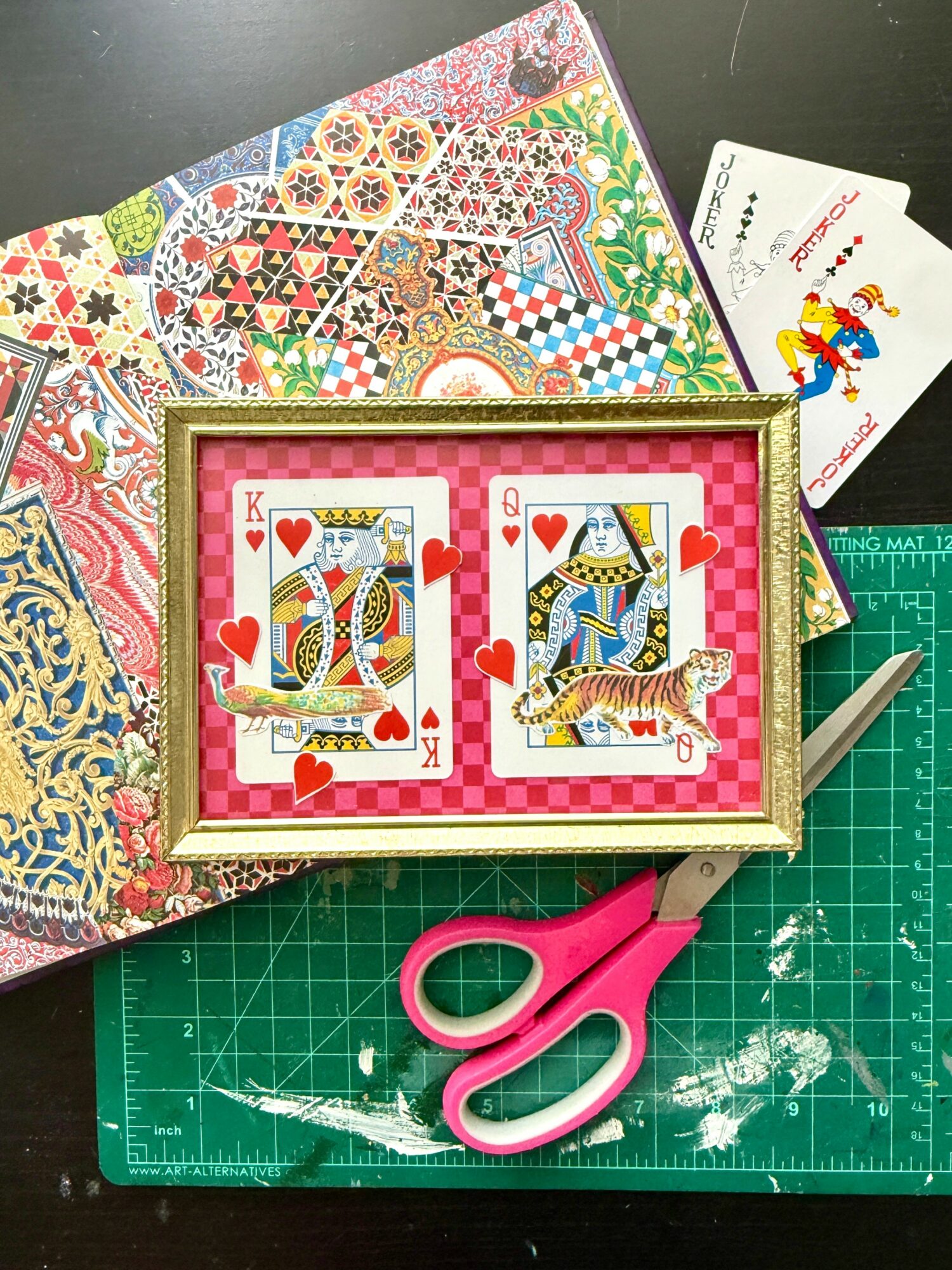 Playing cards with king and queen of hearts and a tiger, on a green cutting mat with scissors and patterned paper.