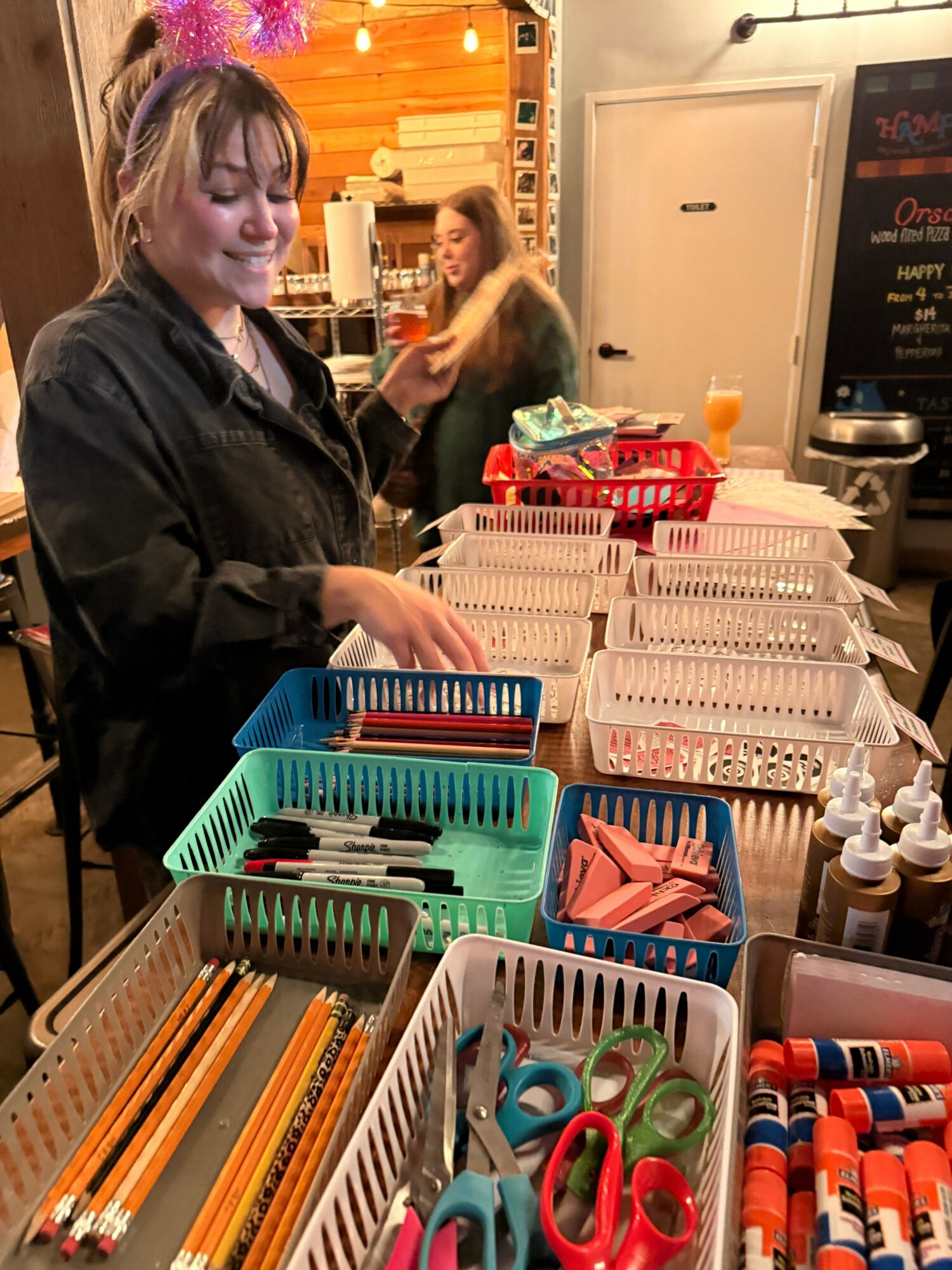 Woman with blonde hair and a black jacket sitting at a table with school supplies, including scissors, pencils, and paper, in a classroom.