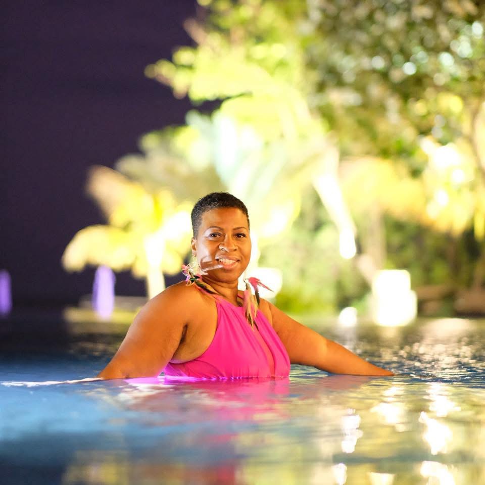 Woman in pink top smiling in a pool with greenery in the background.