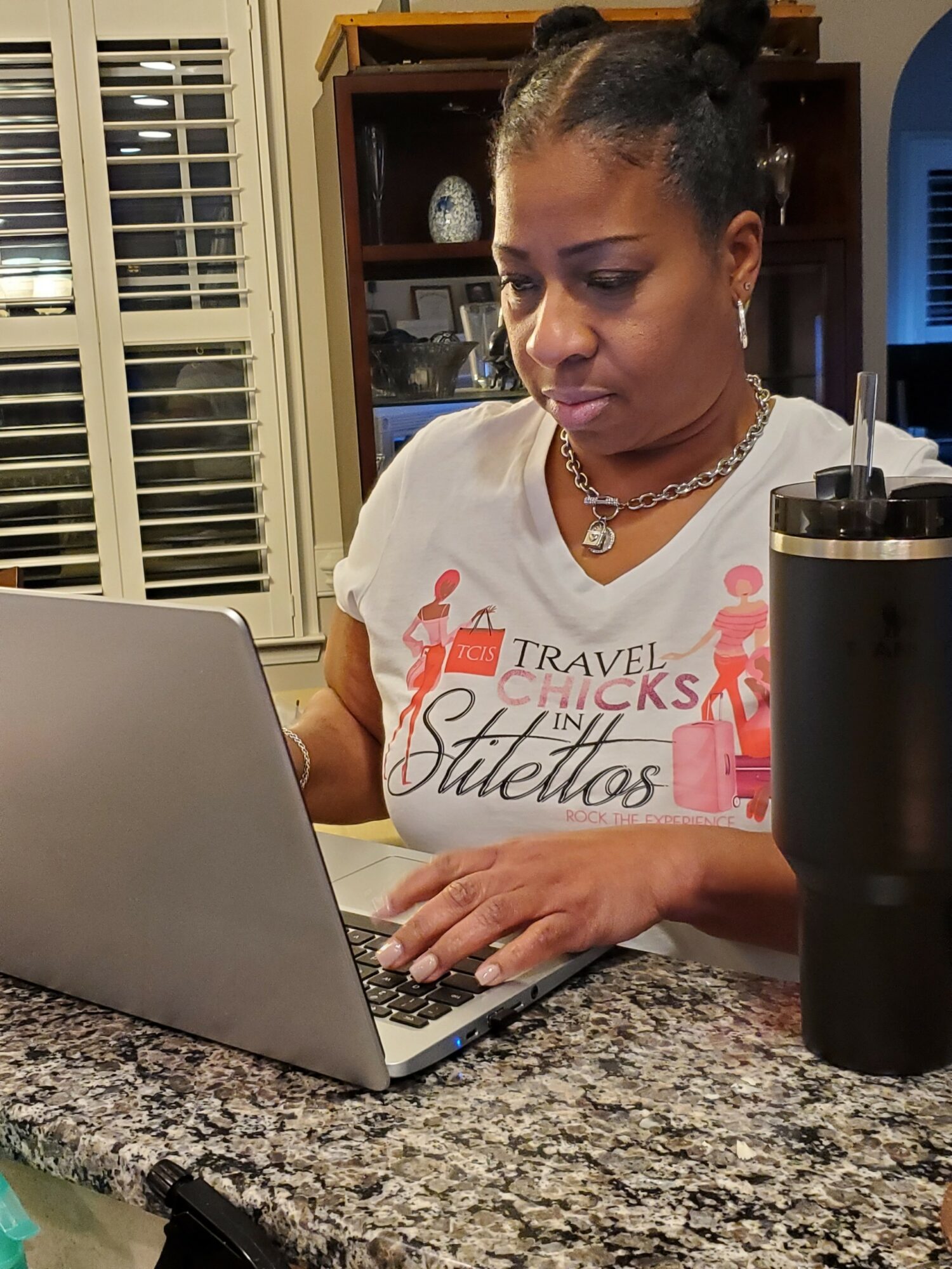 Woman with braided hair working on a laptop at a kitchen counter, with a black tumbler nearby.