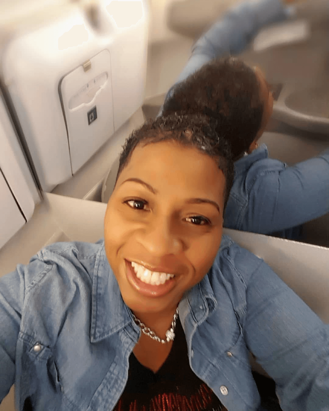 Smiling woman taking selfie on a plane, with a person sitting behind her, wearing a denim jacket.