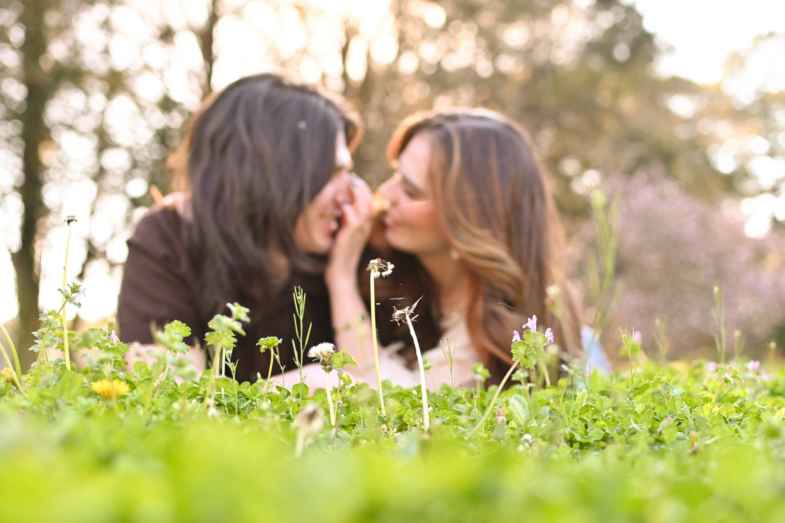 Two women lying in grass, touching noses, outdoors with trees in background, smiling, sunlight filtering through trees.