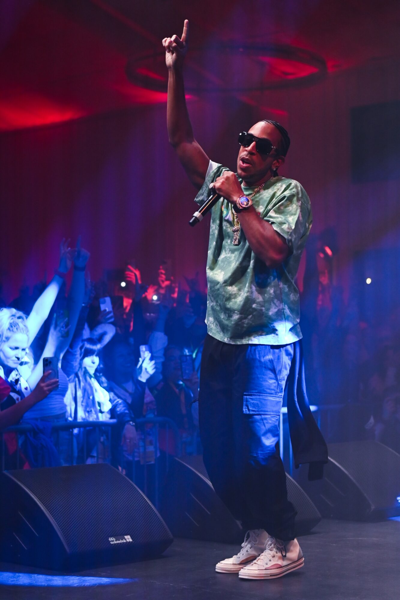 Performer on stage with microphone, raising one arm, wearing sunglasses, patterned shirt, and sneakers, audience in background.