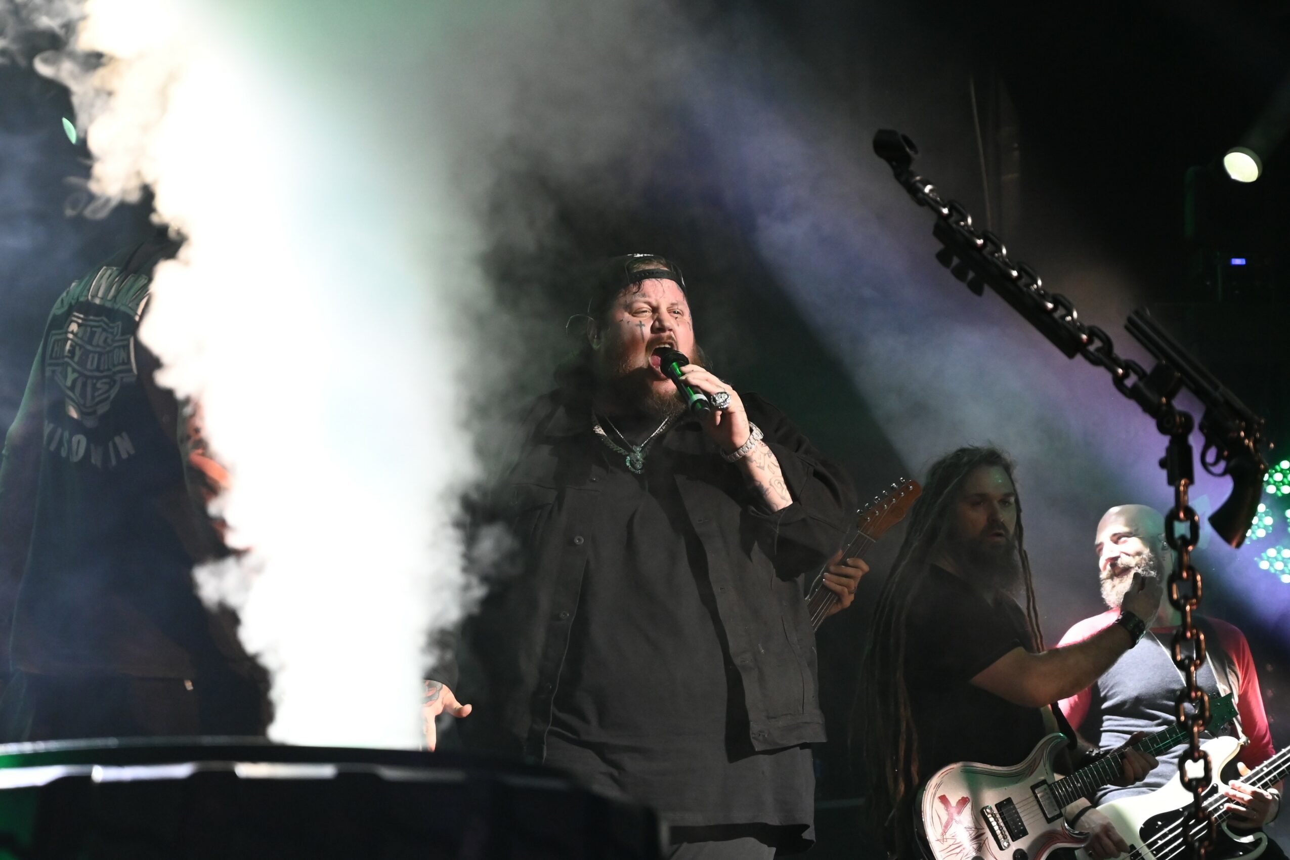 Person singing into microphone on stage with smoke and band members playing guitars, stage lighting visible.