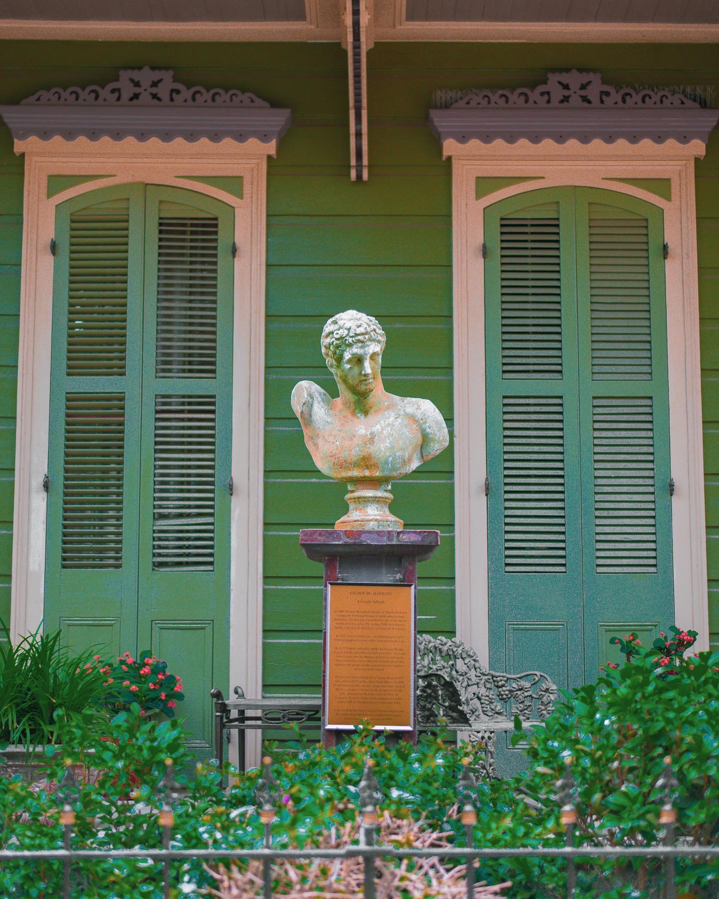 Bust sculpture on pedestal in front of green building with shutters, surrounded by plants and flowers.