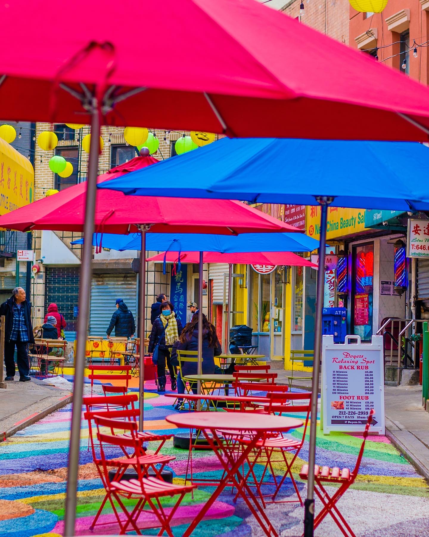 Outdoor seating with red tables and chairs under colorful umbrellas on a city street, with shops and pedestrians in the background.