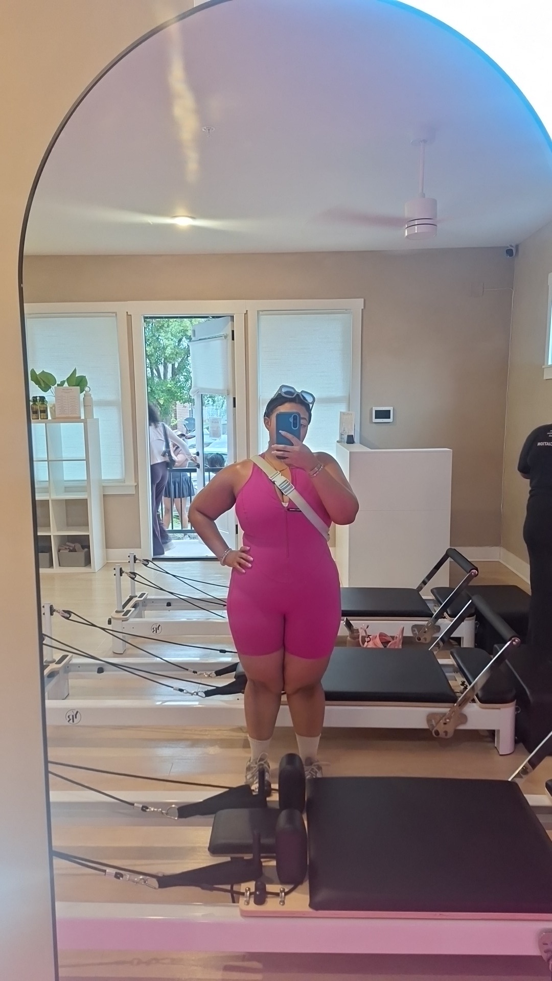 Person taking a mirror selfie in a room with exercise equipment, wearing a pink outfit, holding a phone, with a window in the background.