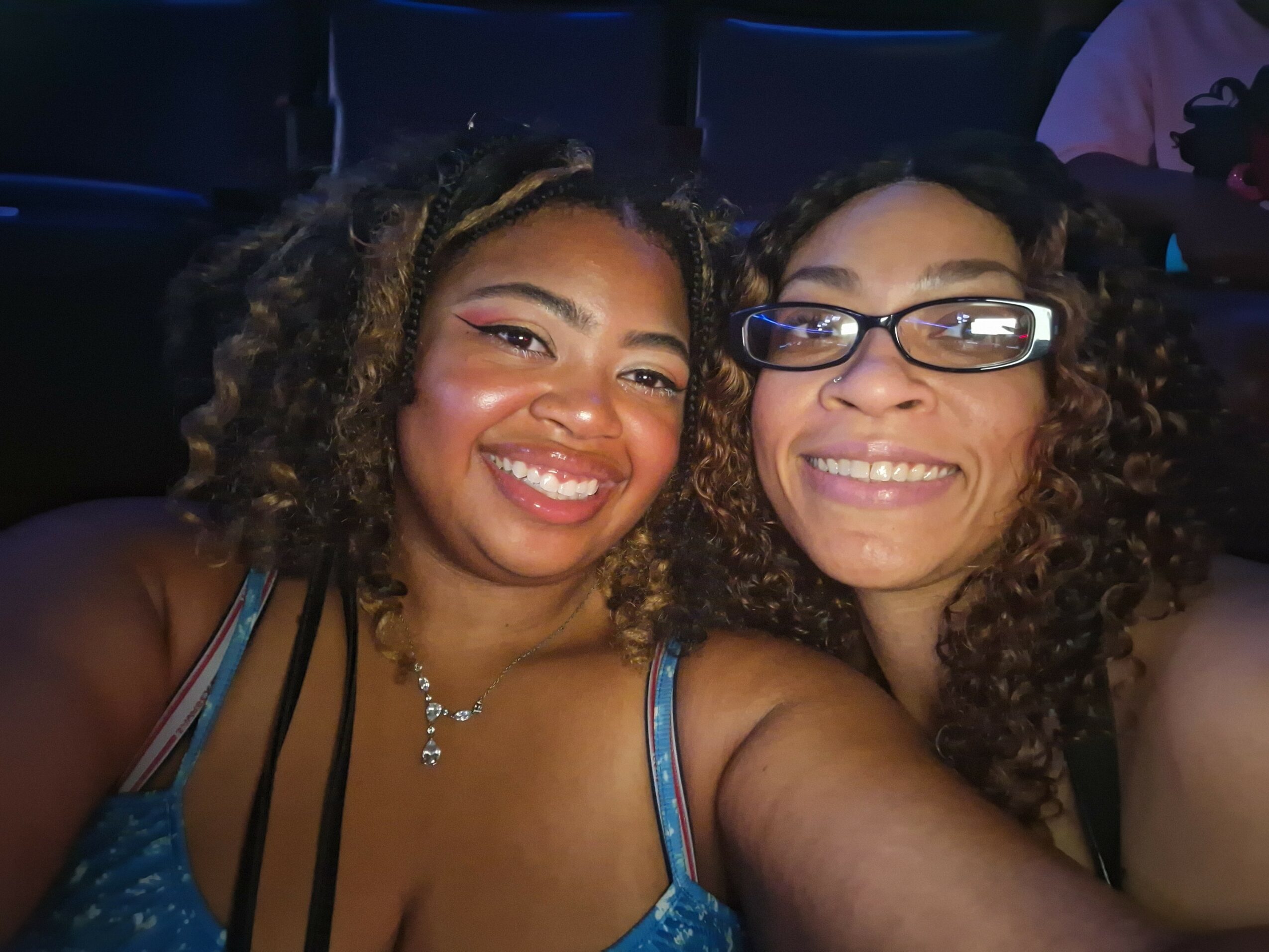 Two women smiling and taking a selfie together, one wearing glasses and the other with curly hair, in a dark setting.