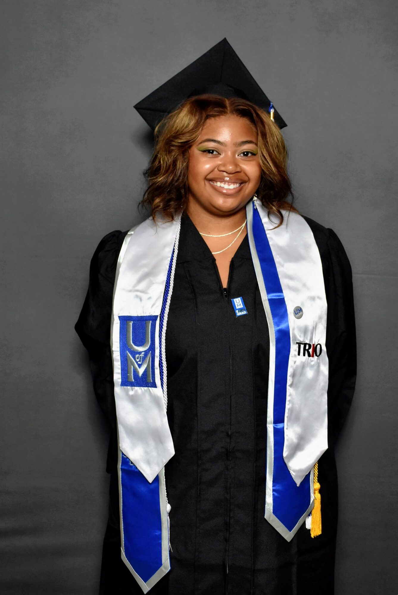 Young woman in graduation cap and gown smiling, standing against a gray background.