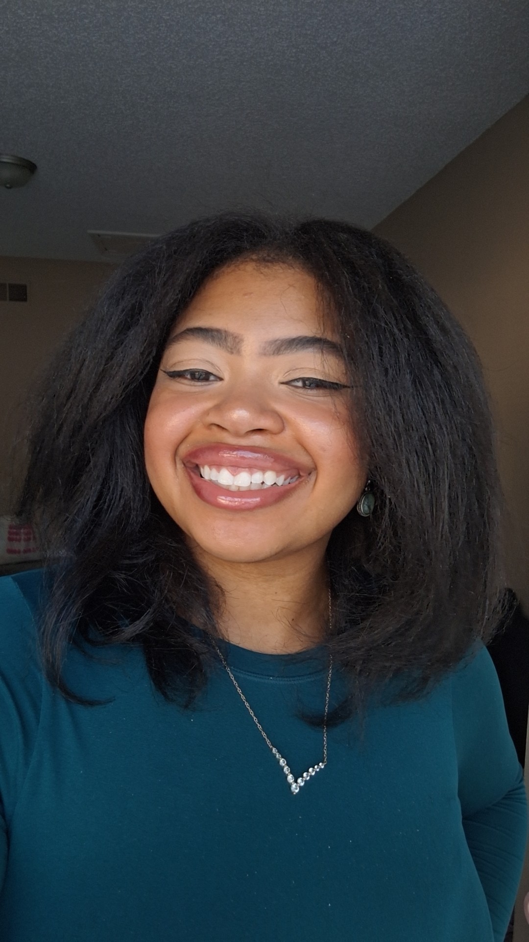 Smiling woman with shoulder-length black hair, wearing a teal top and necklace, indoors.