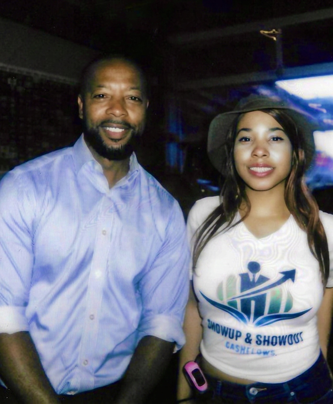 Two people smiling indoors, one man in a light blue shirt and one woman in a white shirt with a logo, wearing a hat.