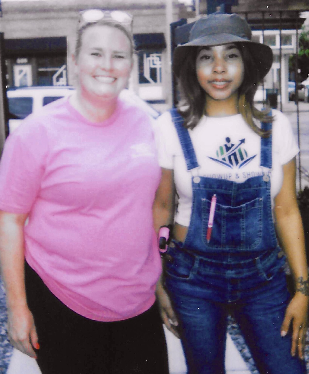 Two women standing outdoors, smiling, one in a pink shirt and the other in overalls and a hat.