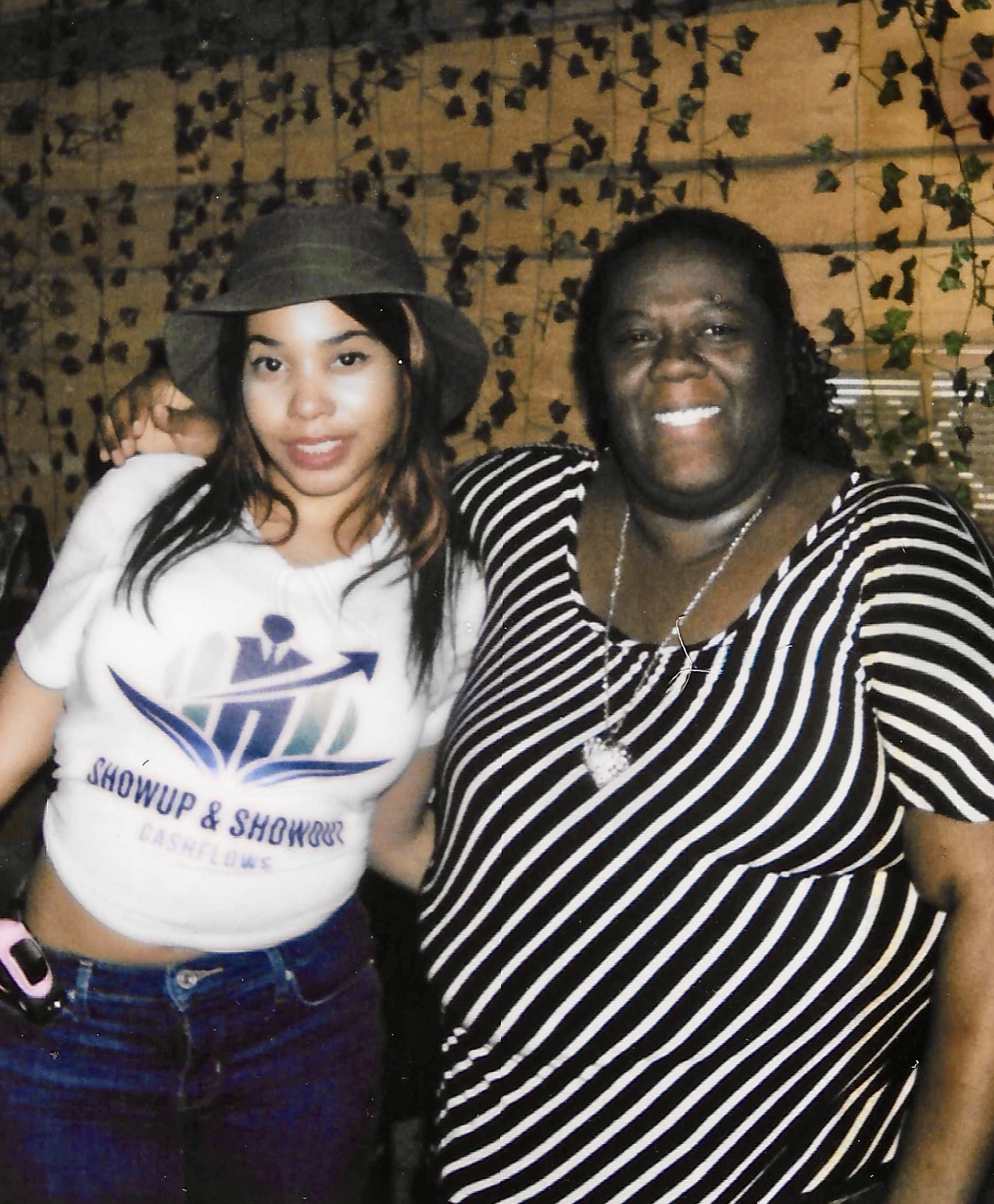 Two women smiling, one wearing a hat and a white shirt, the other in a striped top, standing outdoors with a wooden background and greenery.