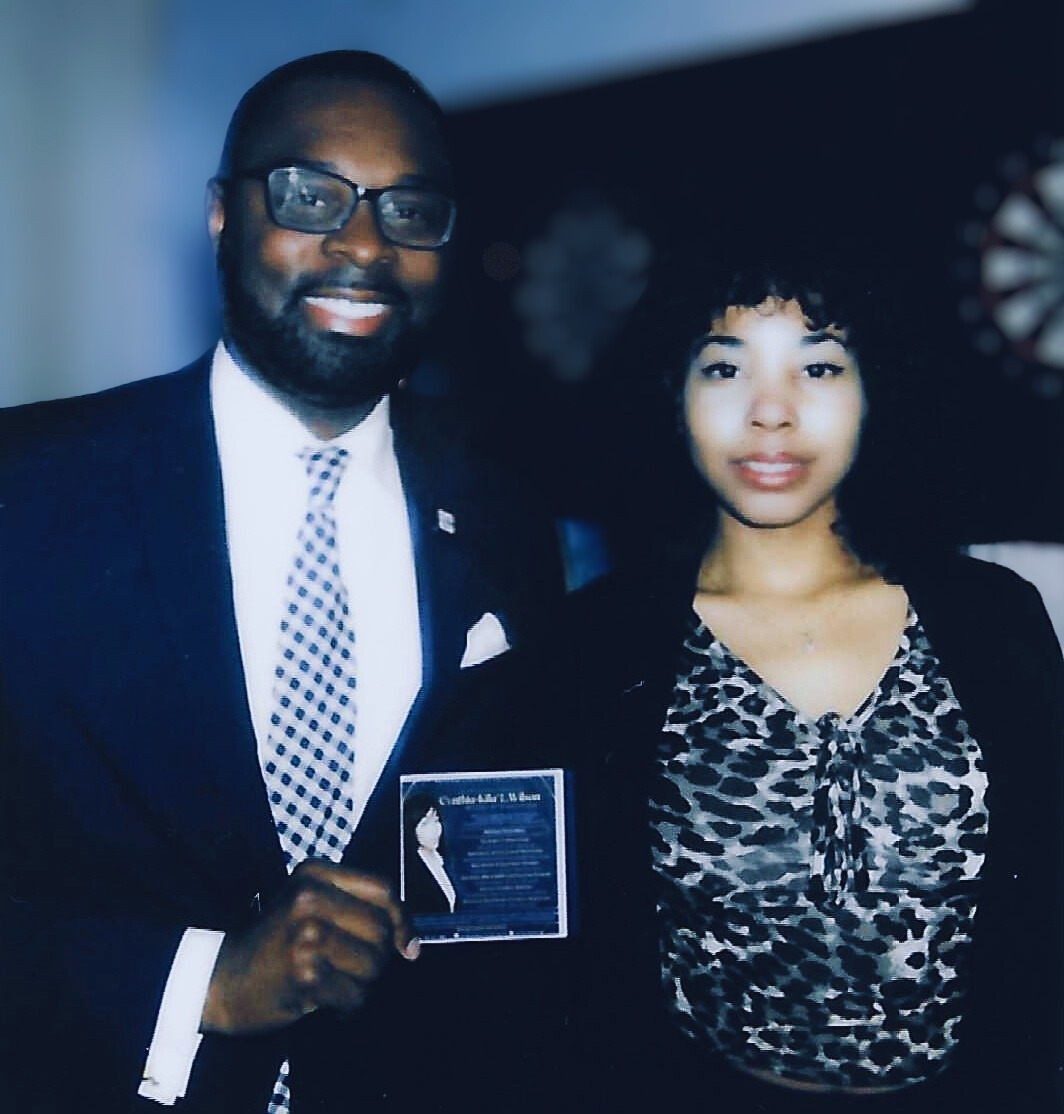 Man in suit and glasses holding an award, woman with curly hair and patterned top standing beside him, dark background.