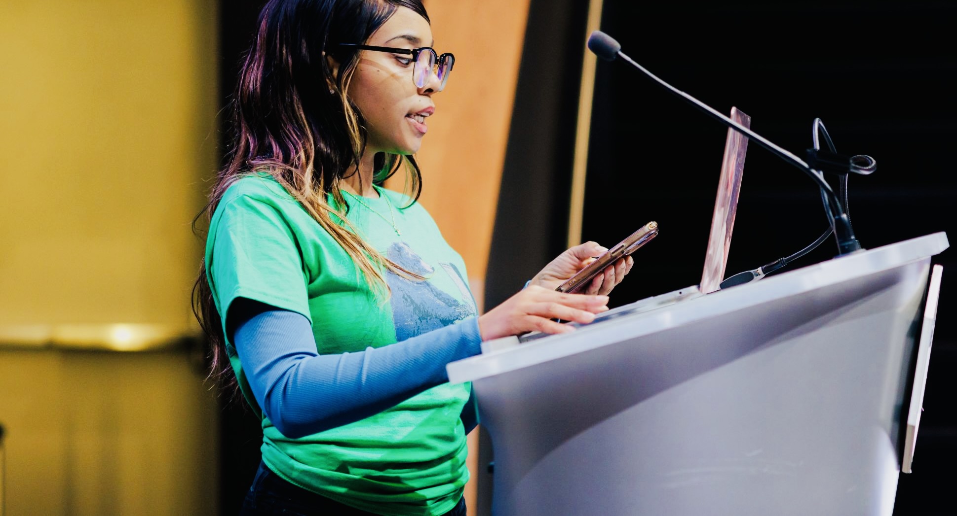 Woman speaking at a podium, holding a phone, wearing glasses and a green shirt, with a microphone in front.