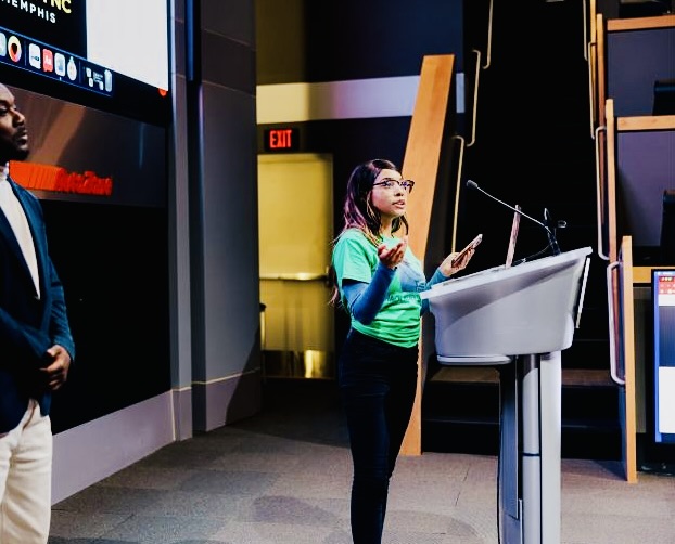 Woman speaking at a podium in a studio or conference room, with a man standing nearby, background includes screens and exit sign.
