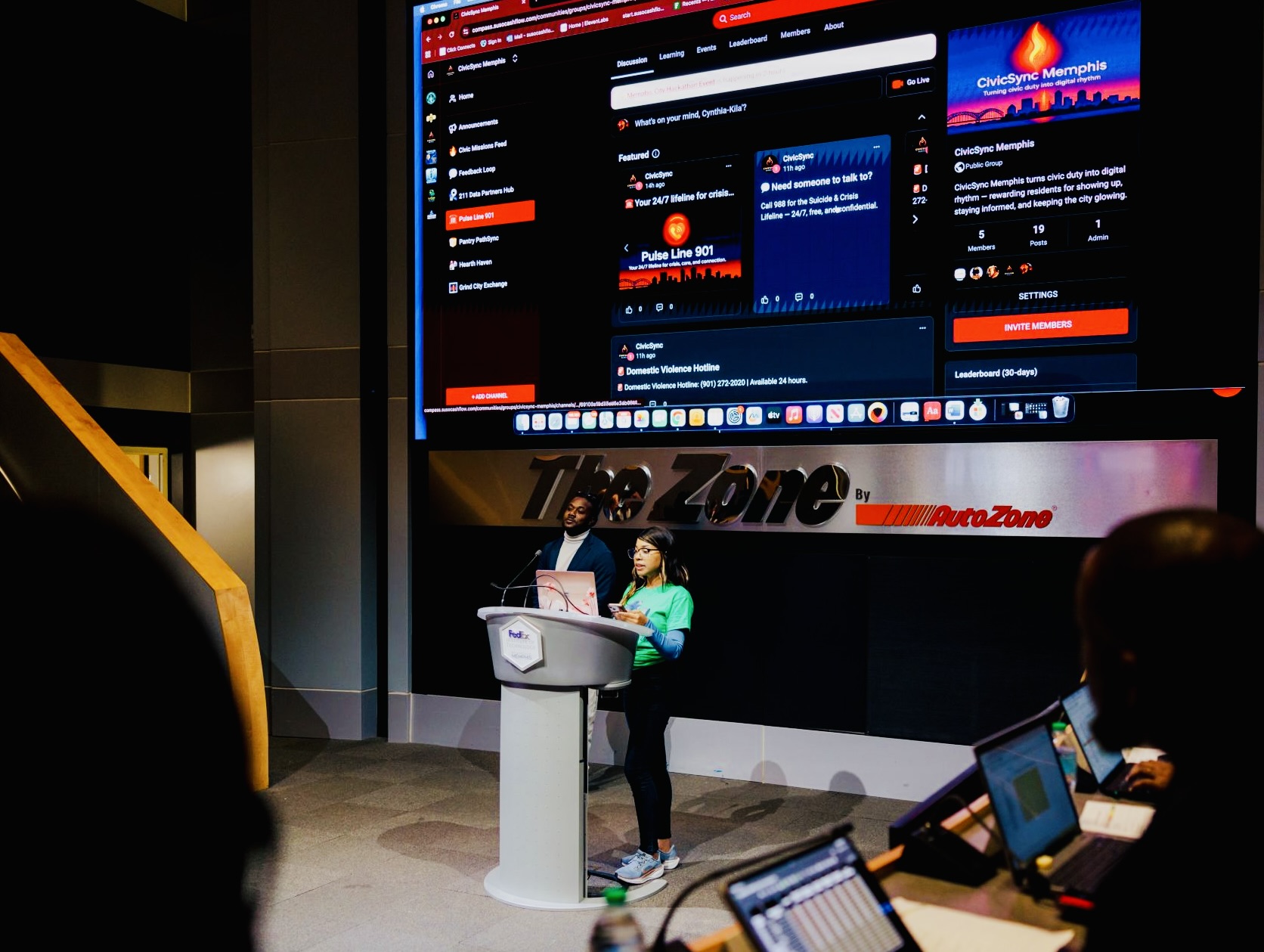 Two people stand at a podium in front of a large screen displaying social media content, with a person working on a laptop nearby.