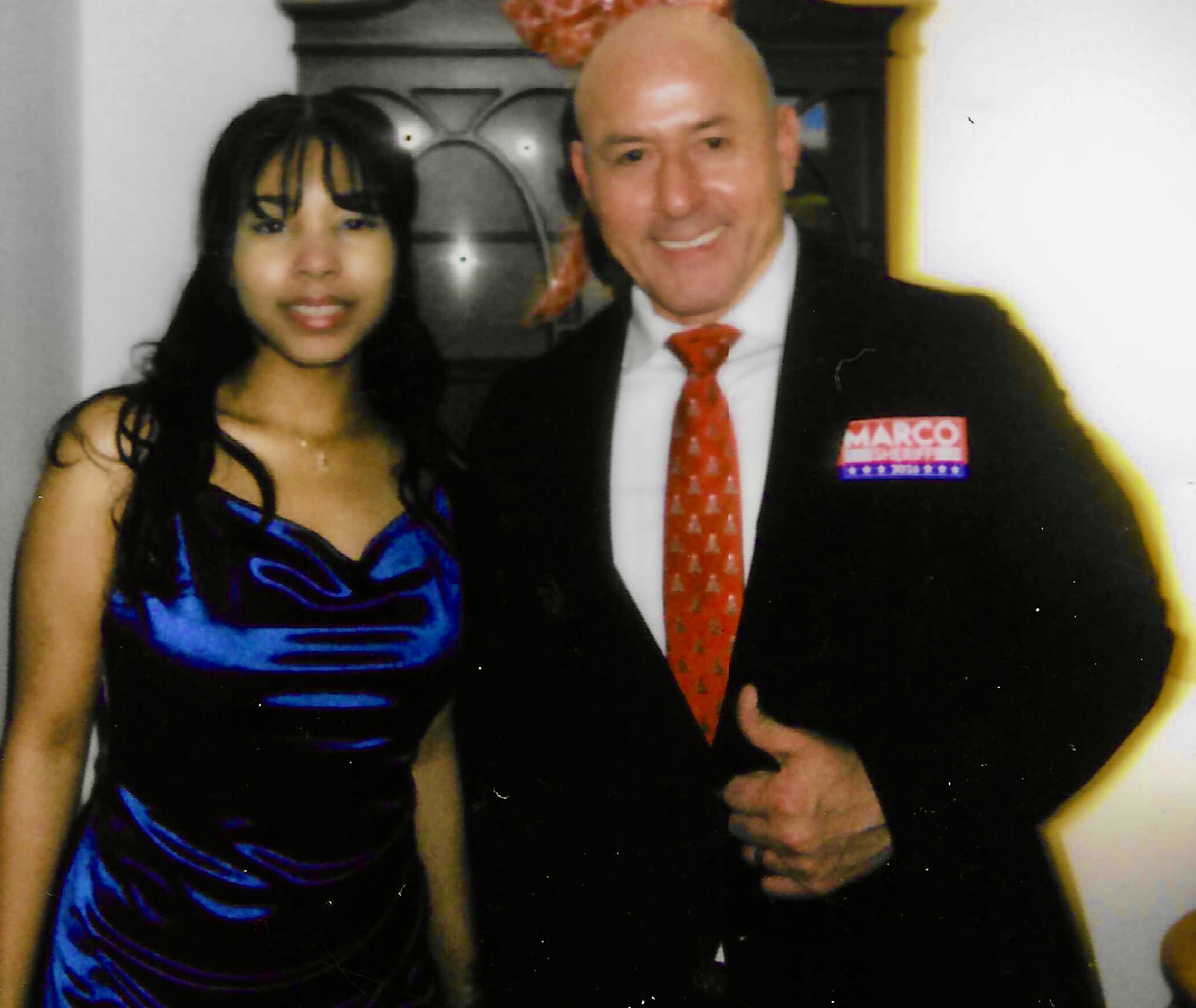 A woman in a blue dress and a man in a suit with a red tie stand together indoors.
