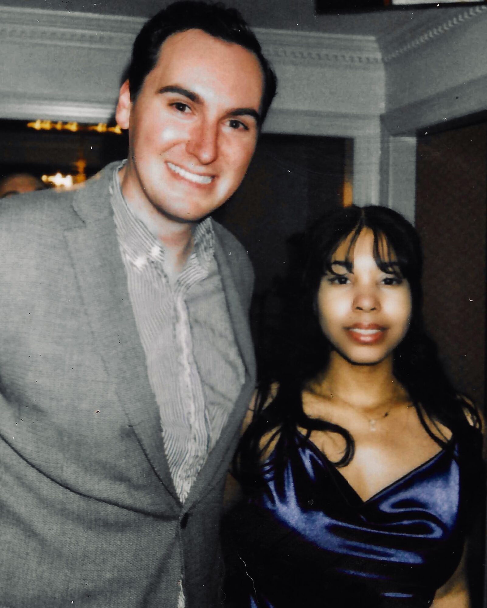 A smiling man and woman pose together indoors, with the man wearing a light gray suit and the woman in a dark blue dress.