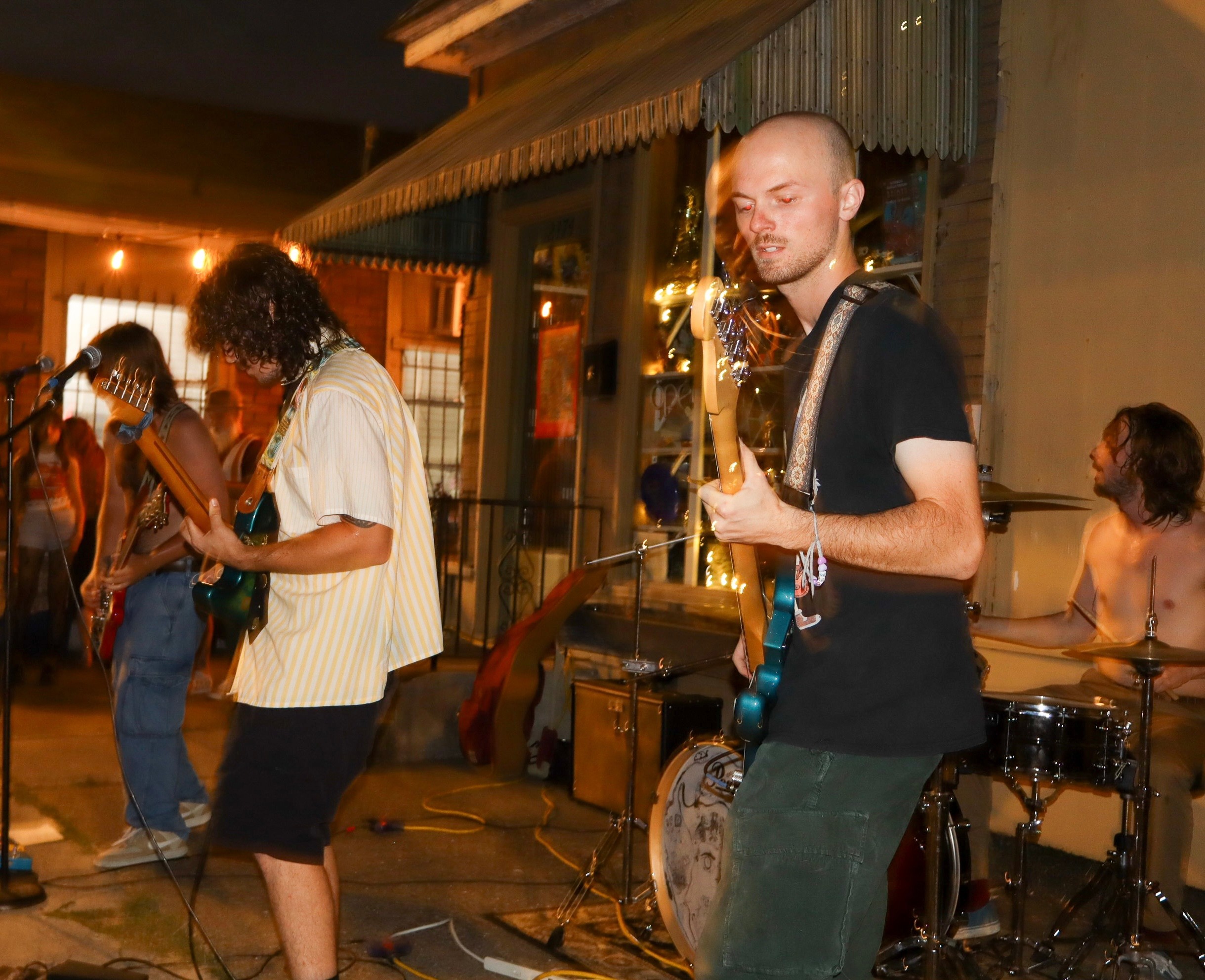 Band performing live with a guitarist, bassist, and drummer in a cozy indoor setting, audience visible in background.