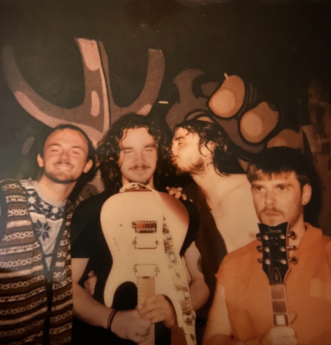 Four men holding guitars, standing in front of a dark background with abstract shapes, some with facial hair and long hair.