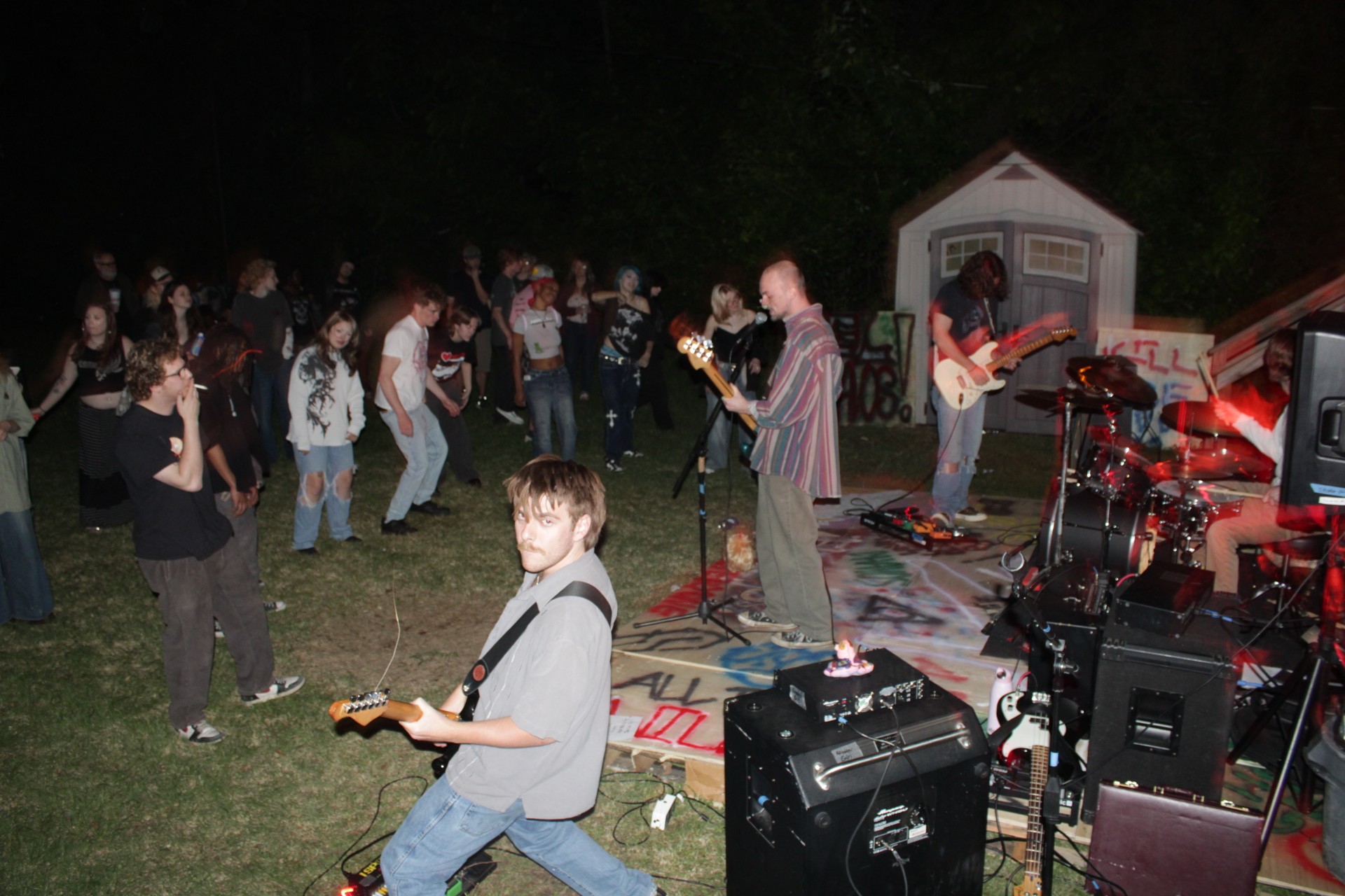 People gather outdoors at night, watching a band perform on a small stage with musical instruments and equipment.