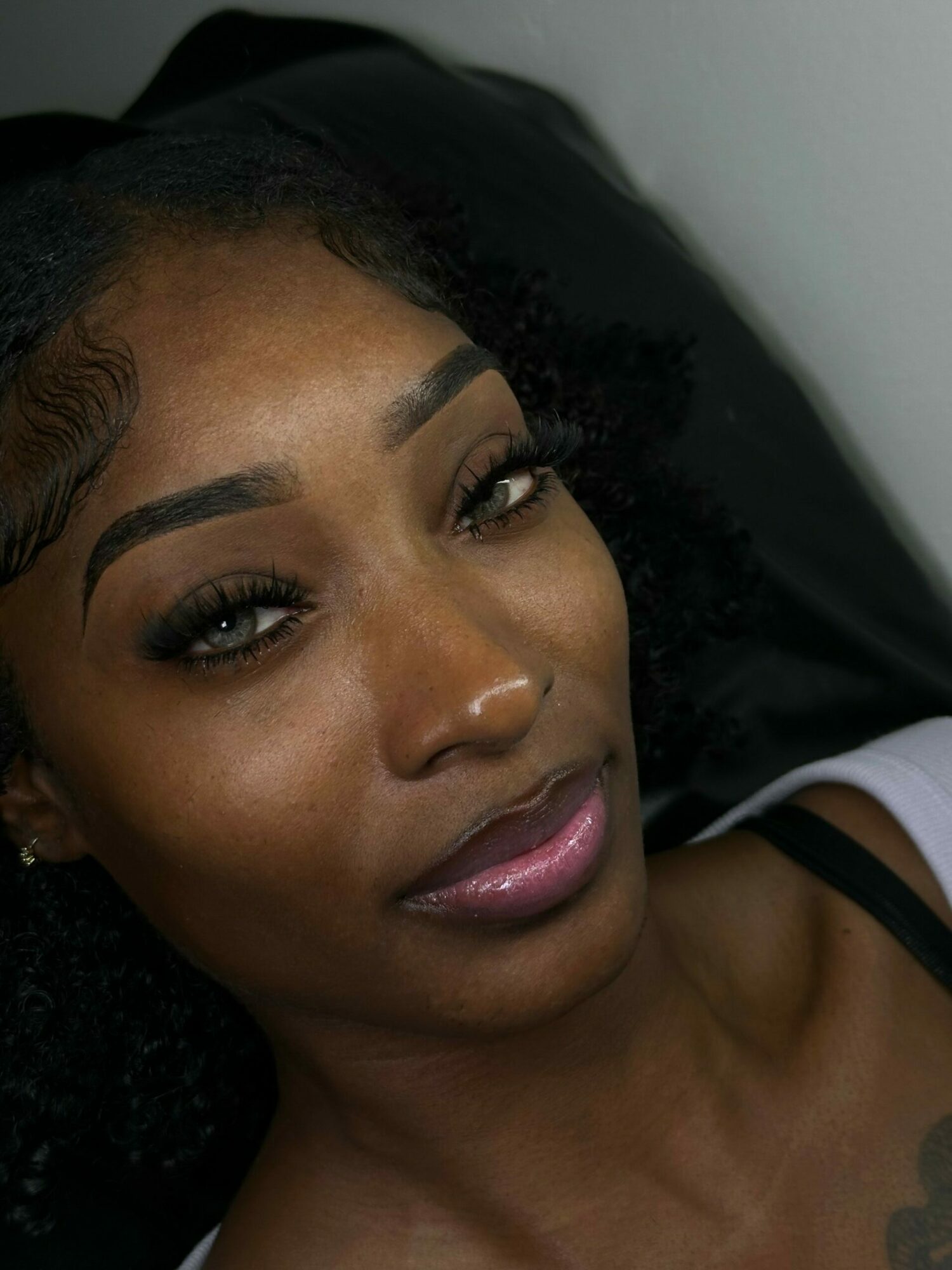 Close-up of a woman's face with makeup, lying down with head tilted back, eyes open, and lips slightly parted.
