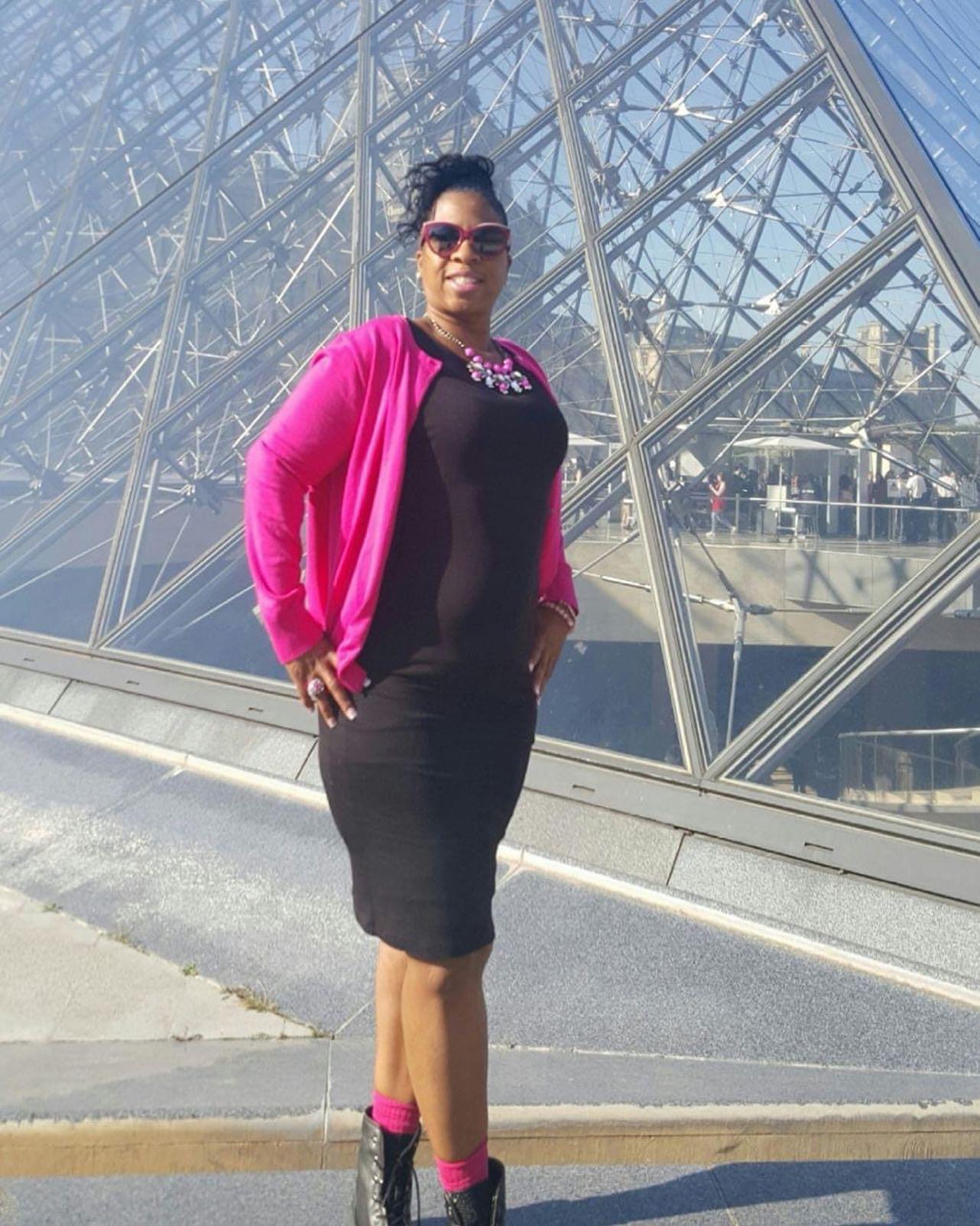 Woman standing outdoors in front of a glass building, wearing sunglasses, a black dress, pink cardigan, and pink socks.