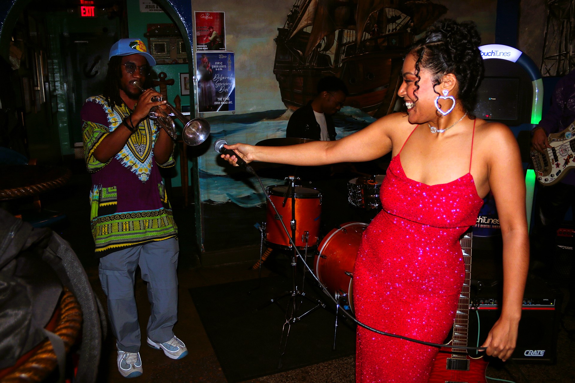 Woman in a red dress singing into a microphone, others playing instruments in a dimly lit room.
