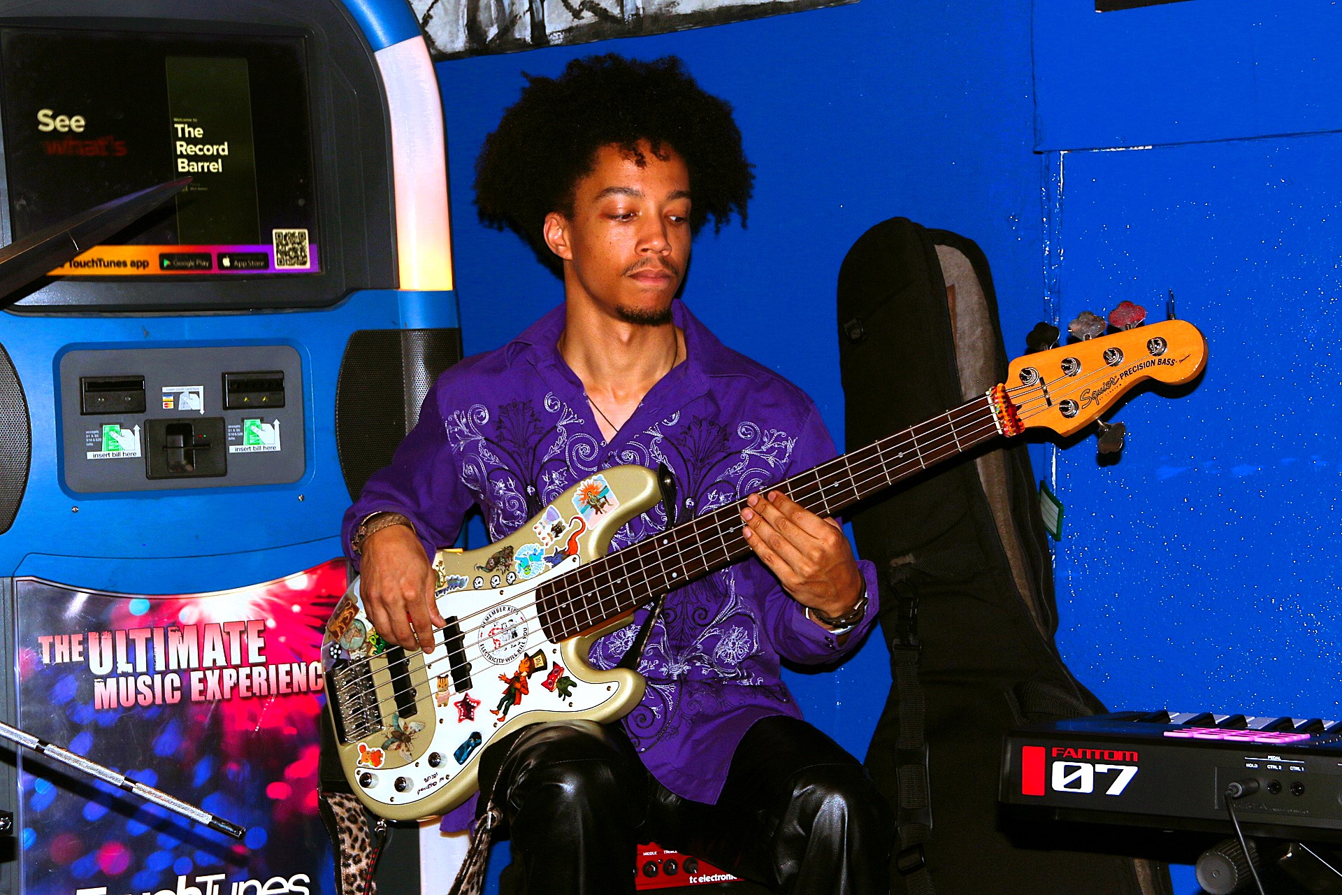 Person with curly hair playing a decorated electric bass guitar in a music arcade setting, blue wall background.