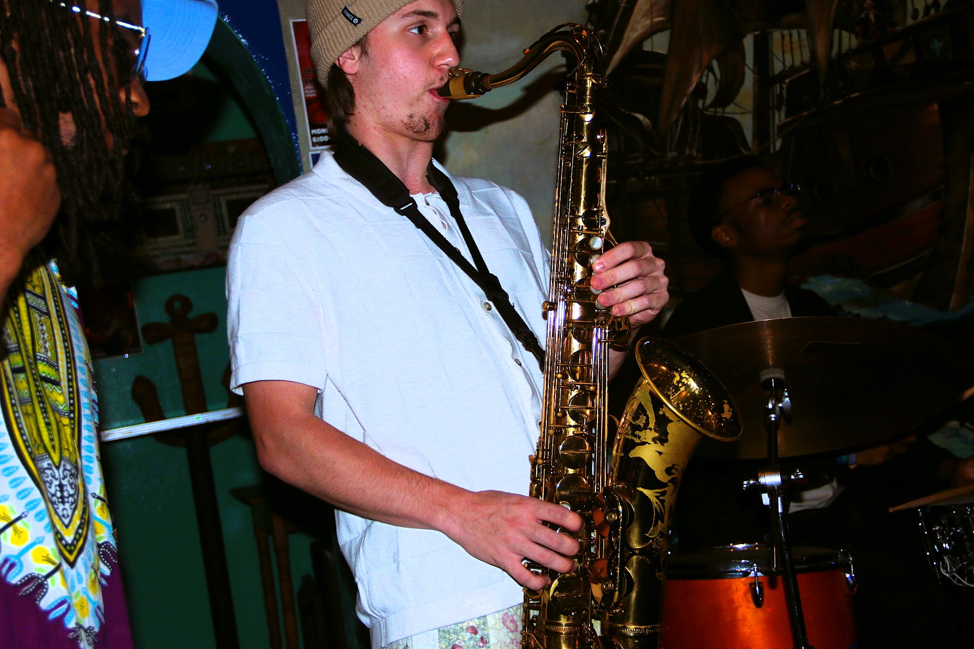 Person playing saxophone in a room with other people and musical instruments.