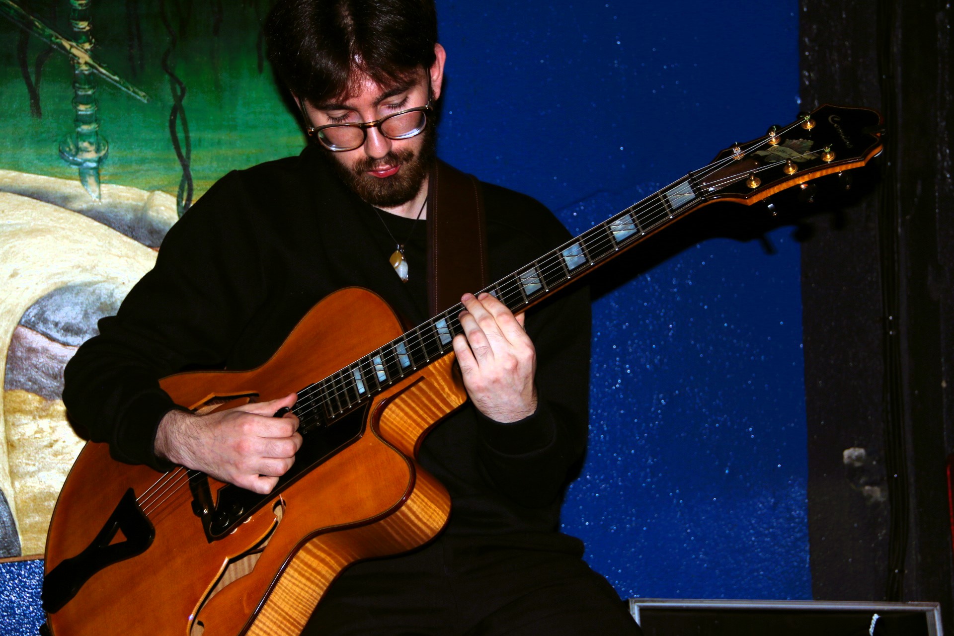 Person playing an acoustic guitar on stage with a colorful background, wearing glasses and a black jacket.
