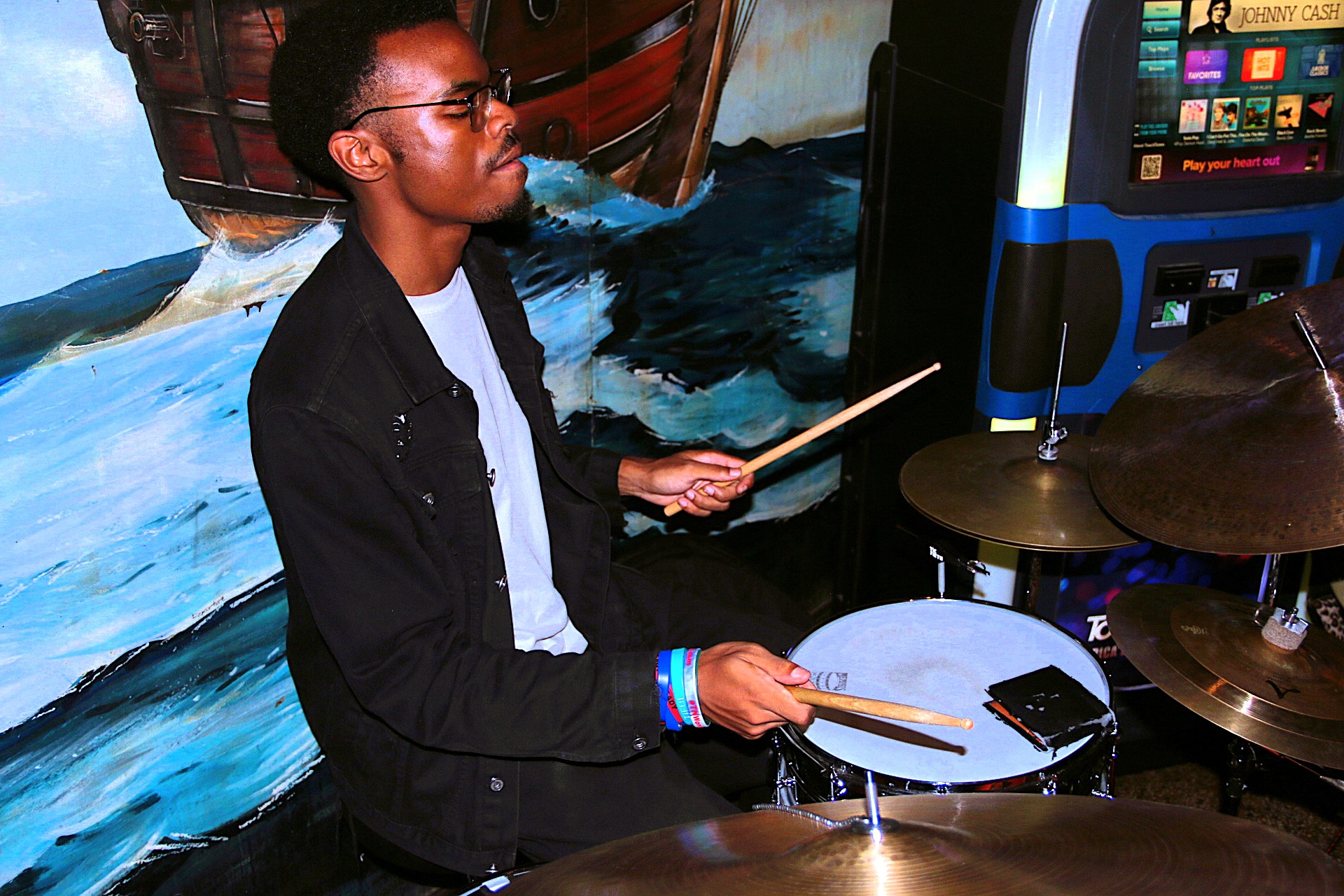 Person playing drums with drumsticks, wearing glasses and a black jacket, in front of a colorful mural background.
