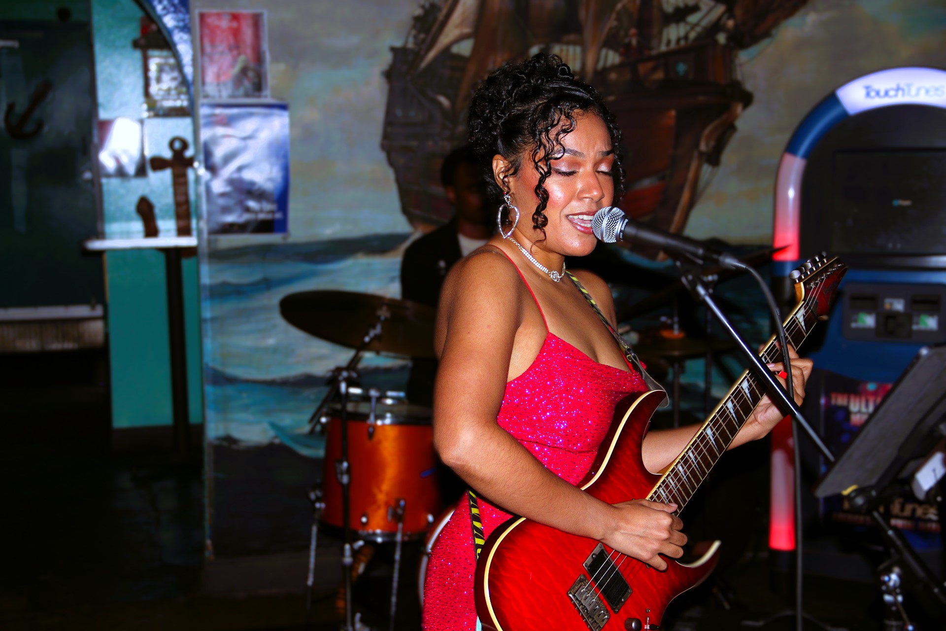 A woman singing into a microphone while playing an electric guitar on stage, with a drum set in the background.