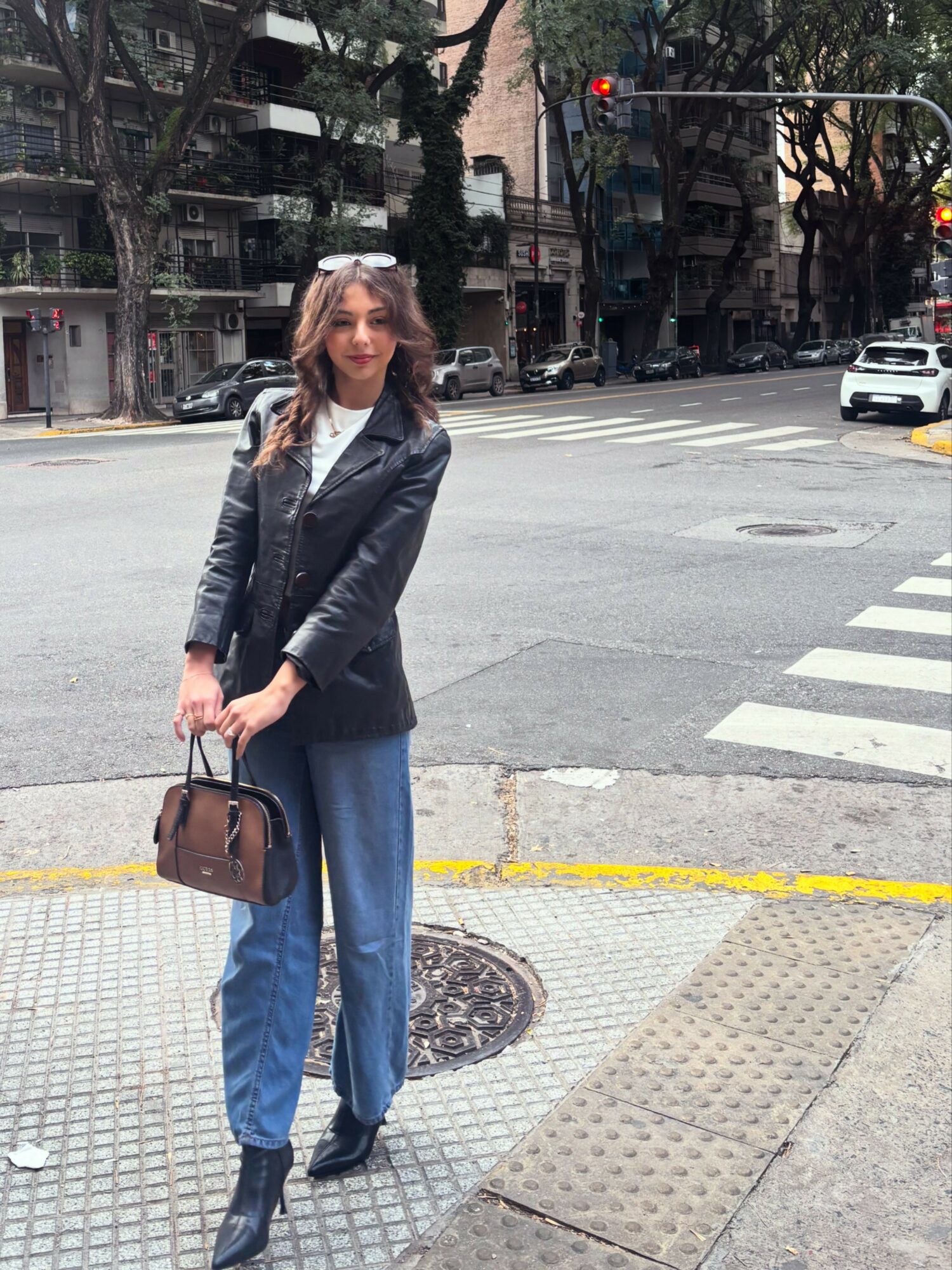 Woman standing on sidewalk holding a brown handbag, wearing a black jacket, blue jeans, and black boots, in an urban street scene.