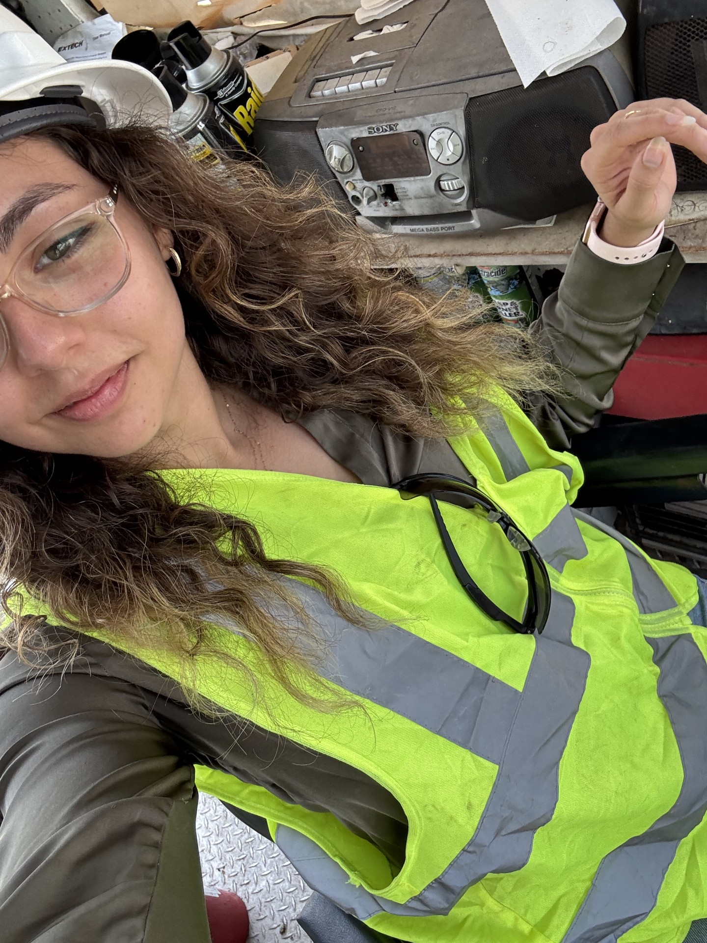 Woman with curly hair wearing safety glasses and a high-visibility vest, sitting in a cluttered workspace.