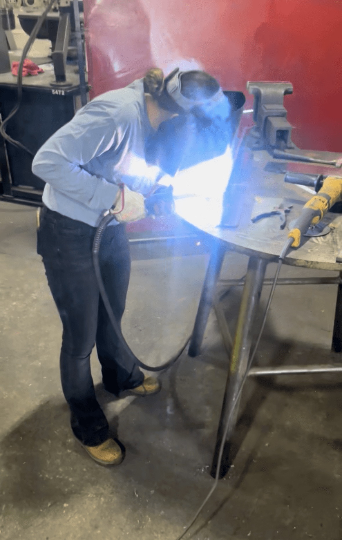 Person welding metal at a workbench, wearing protective gear, sparks and bright light from welding process, workshop setting