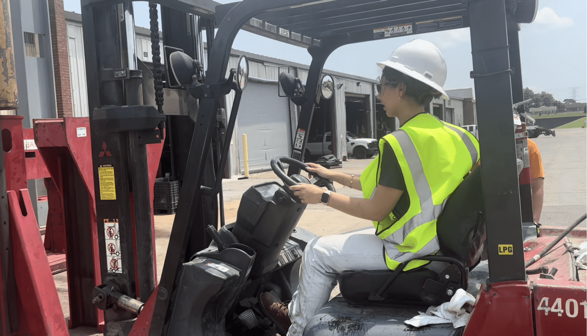 Person wearing a white helmet and yellow safety vest operating a forklift outdoors.