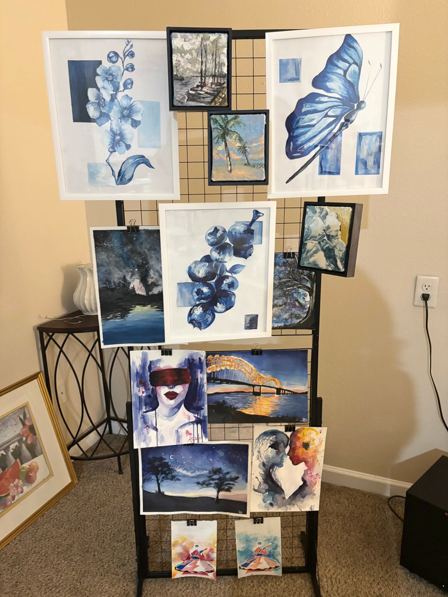 Display stand with various framed artwork, including butterflies, landscapes, and abstract images, arranged on a beige wall and floor.