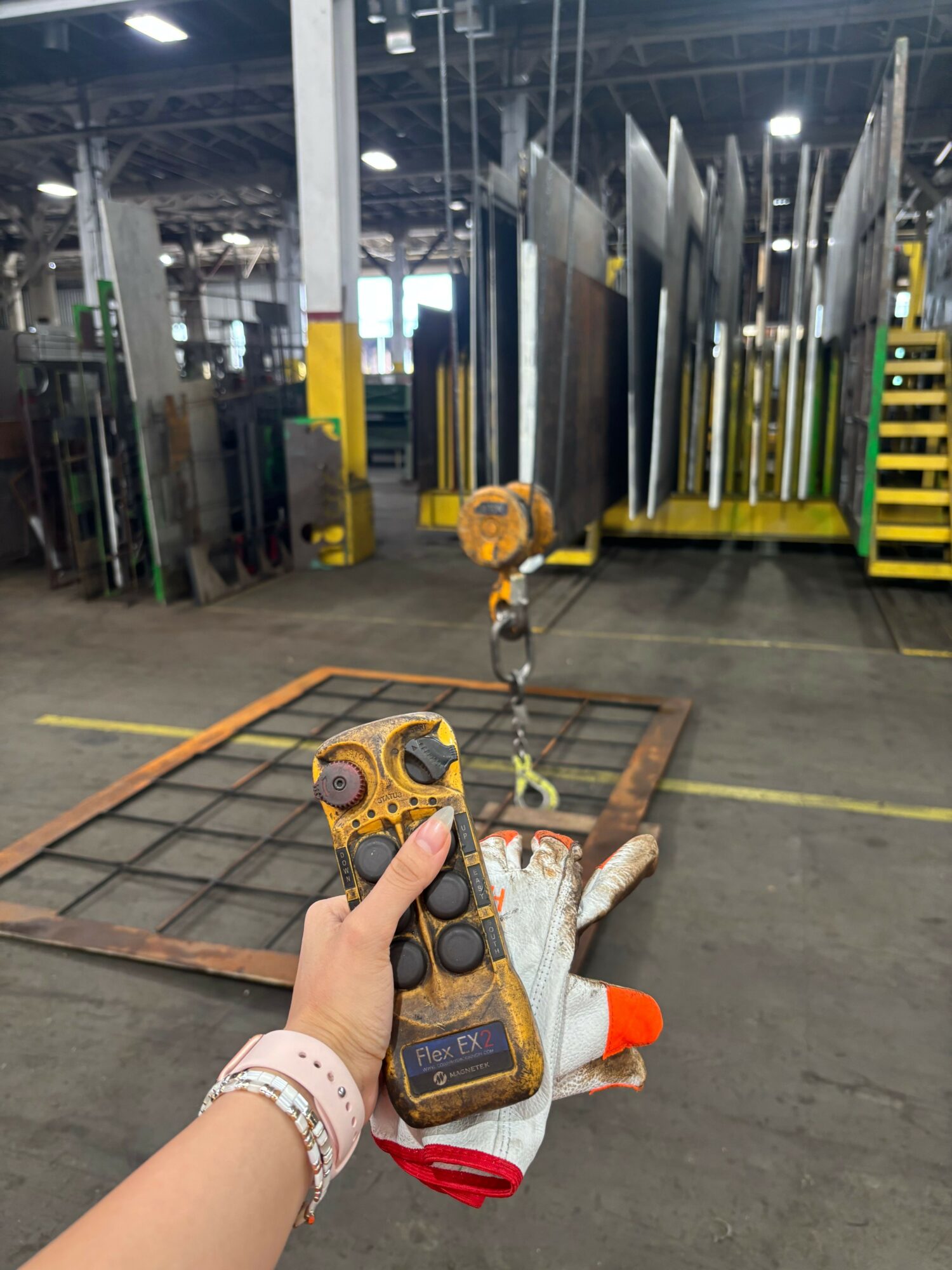 Person holding a remote control in a warehouse with pipes and equipment, with a worker in the background.