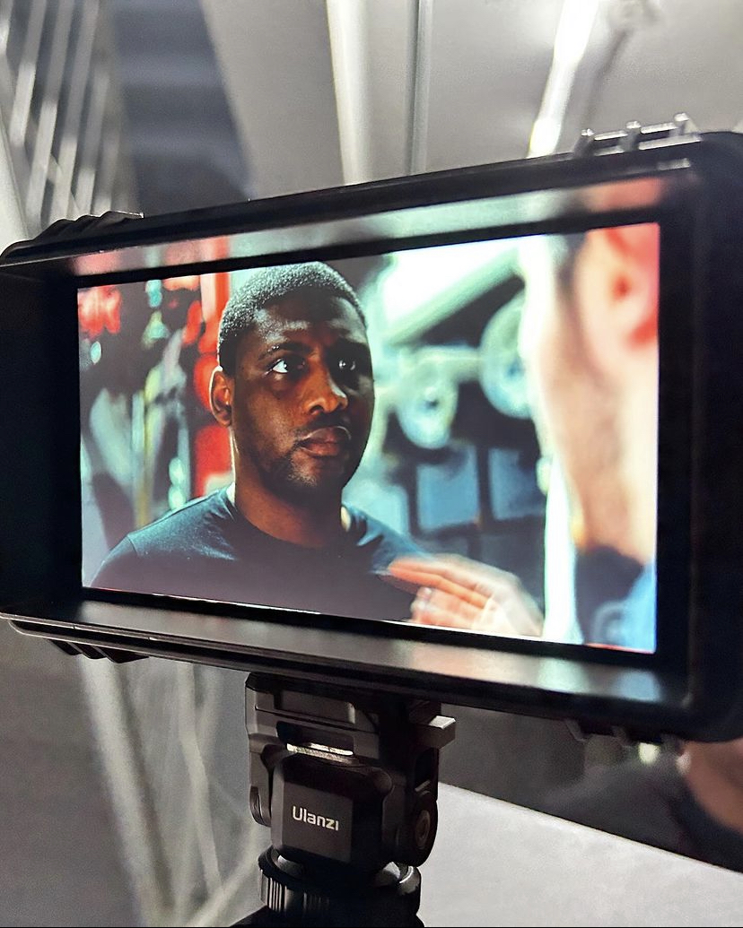 A man with short hair and a beard looks at another person in a gym setting with weights in the background.