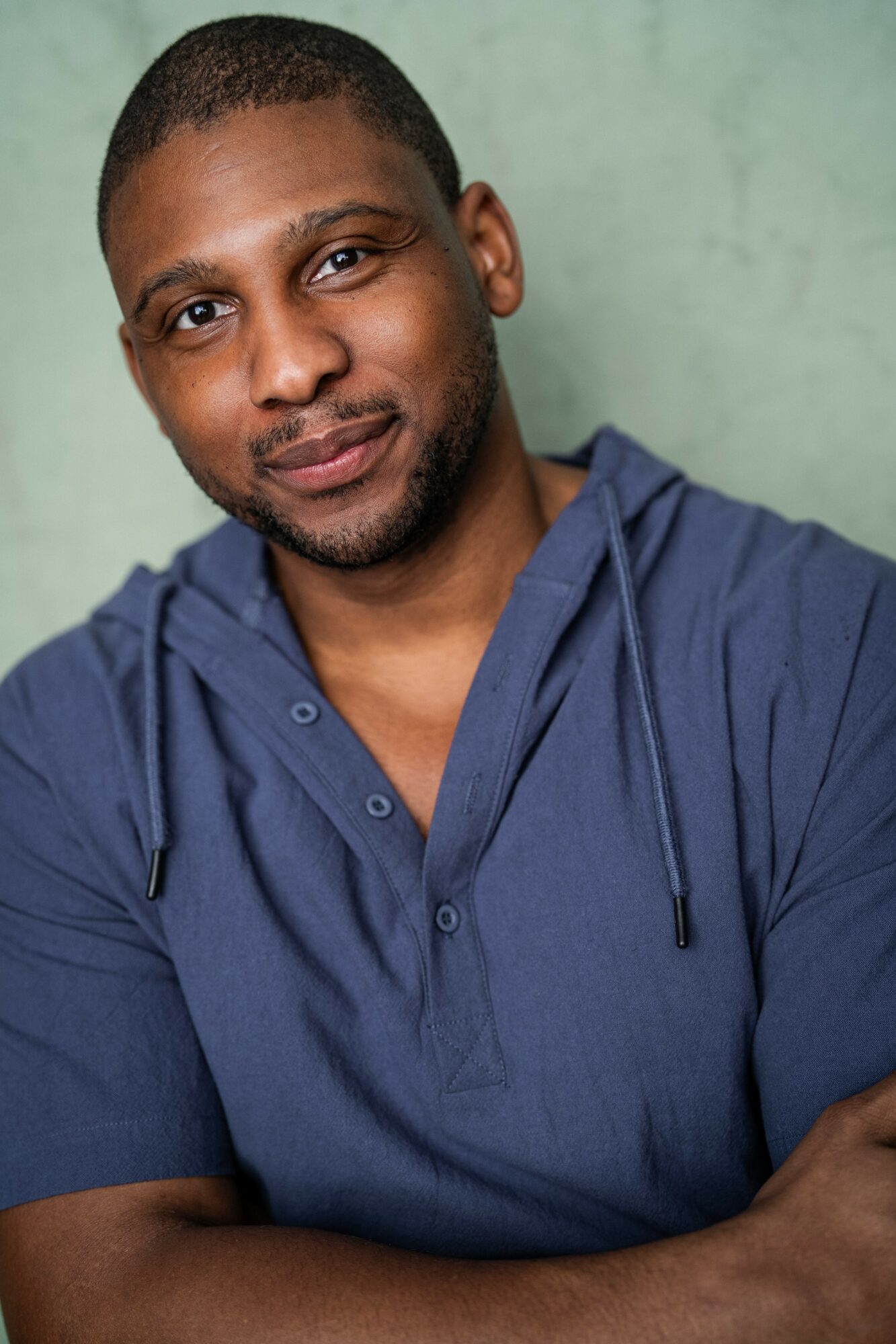 Man with short hair and a beard wearing a blue hoodie, smiling, against a green background.