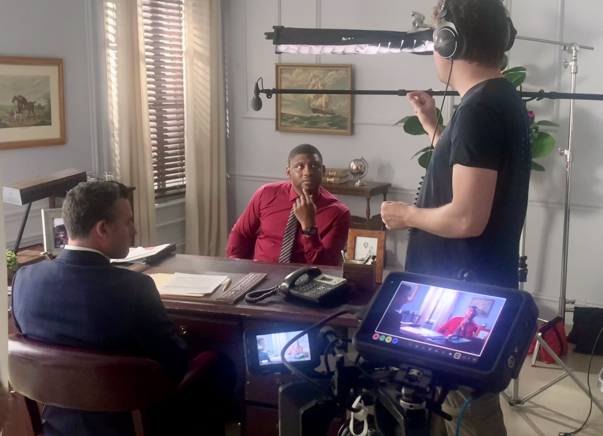 A woman in a red jacket sits at a desk with two men, one in a black shirt and the other in a suit, in a room with window blinds and framed pictures. A camera and crew are filming the scene.
