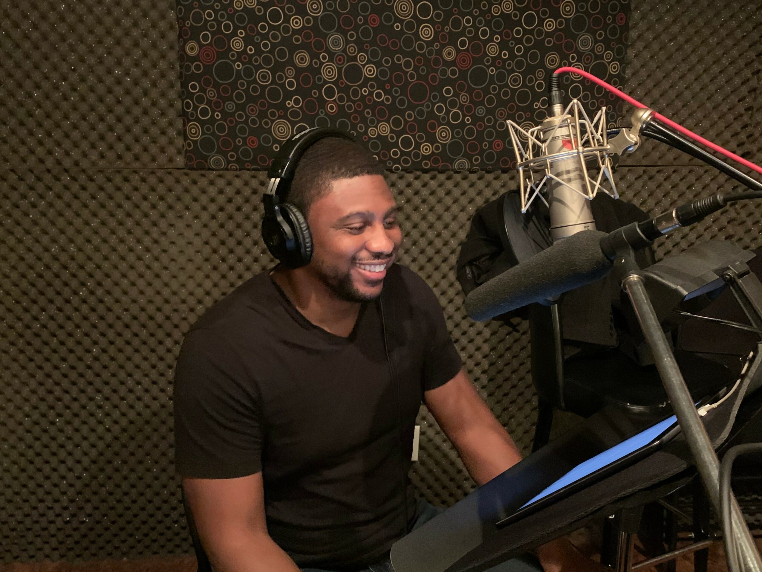 Man with headphones smiling at microphone in recording studio, surrounded by soundproofing panels.