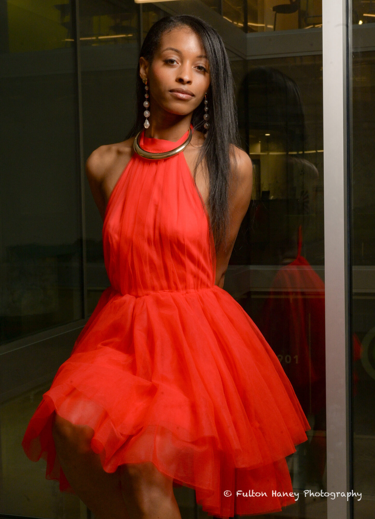 Young woman in a red dress with tulle skirt, standing indoors near glass windows, looking at camera.