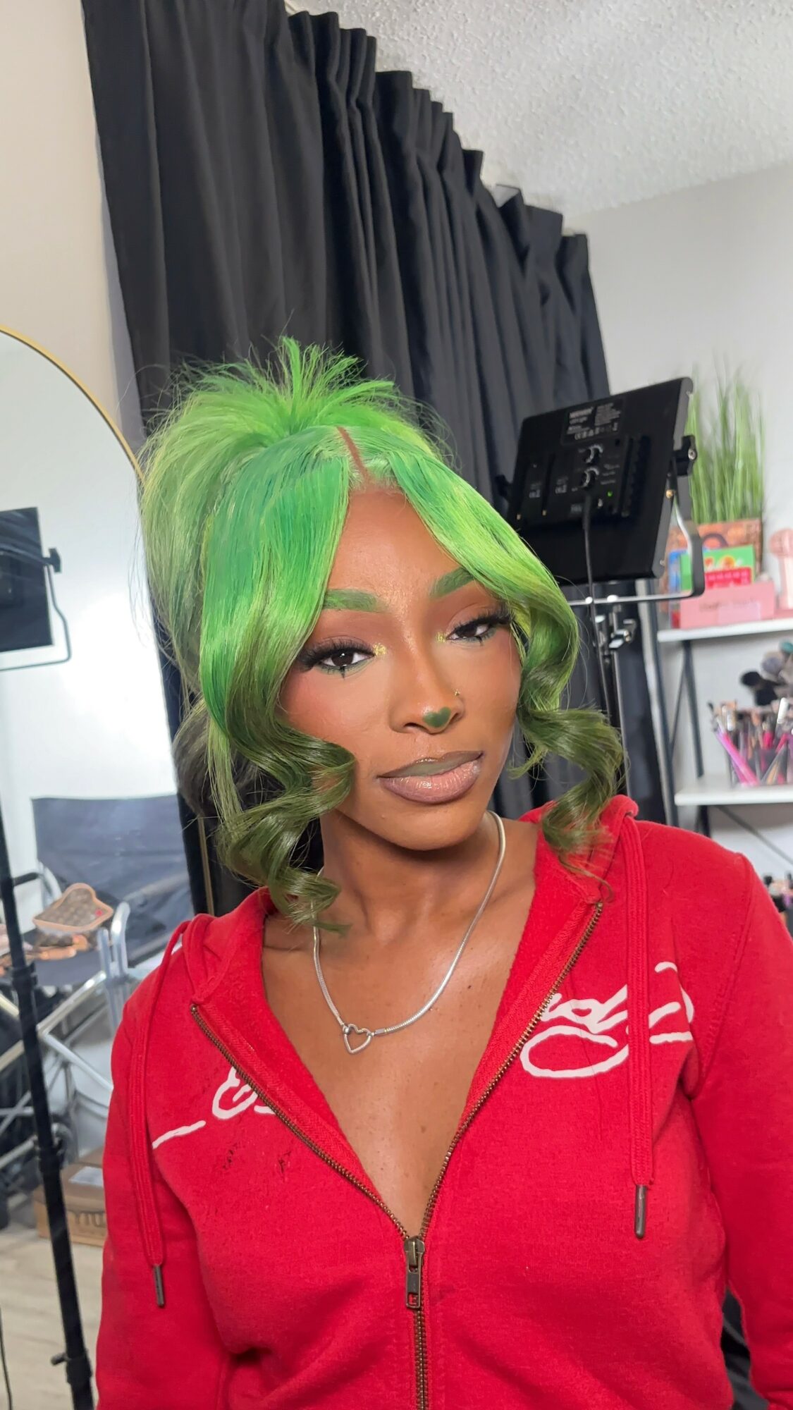 Woman with bright green hair styled in loose waves, wearing a red zip-up hoodie and a necklace, in a room with black curtains and shelves.