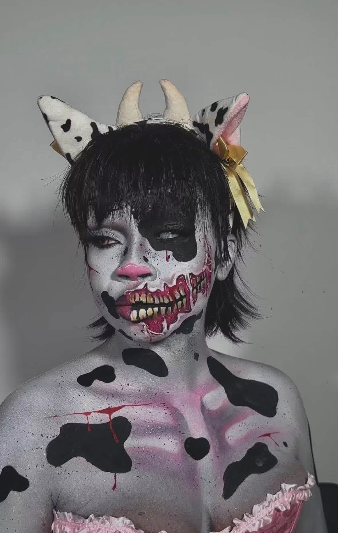 Person with face painted as a cow, wearing a headband with horns and ears, and a yellow bow, against a plain background.