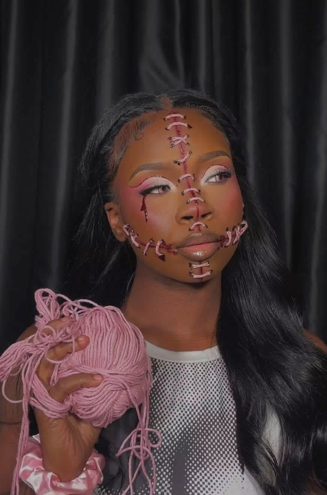 Woman with long black hair and face painted with pink stitches, holding pink yarn, against black curtain background.