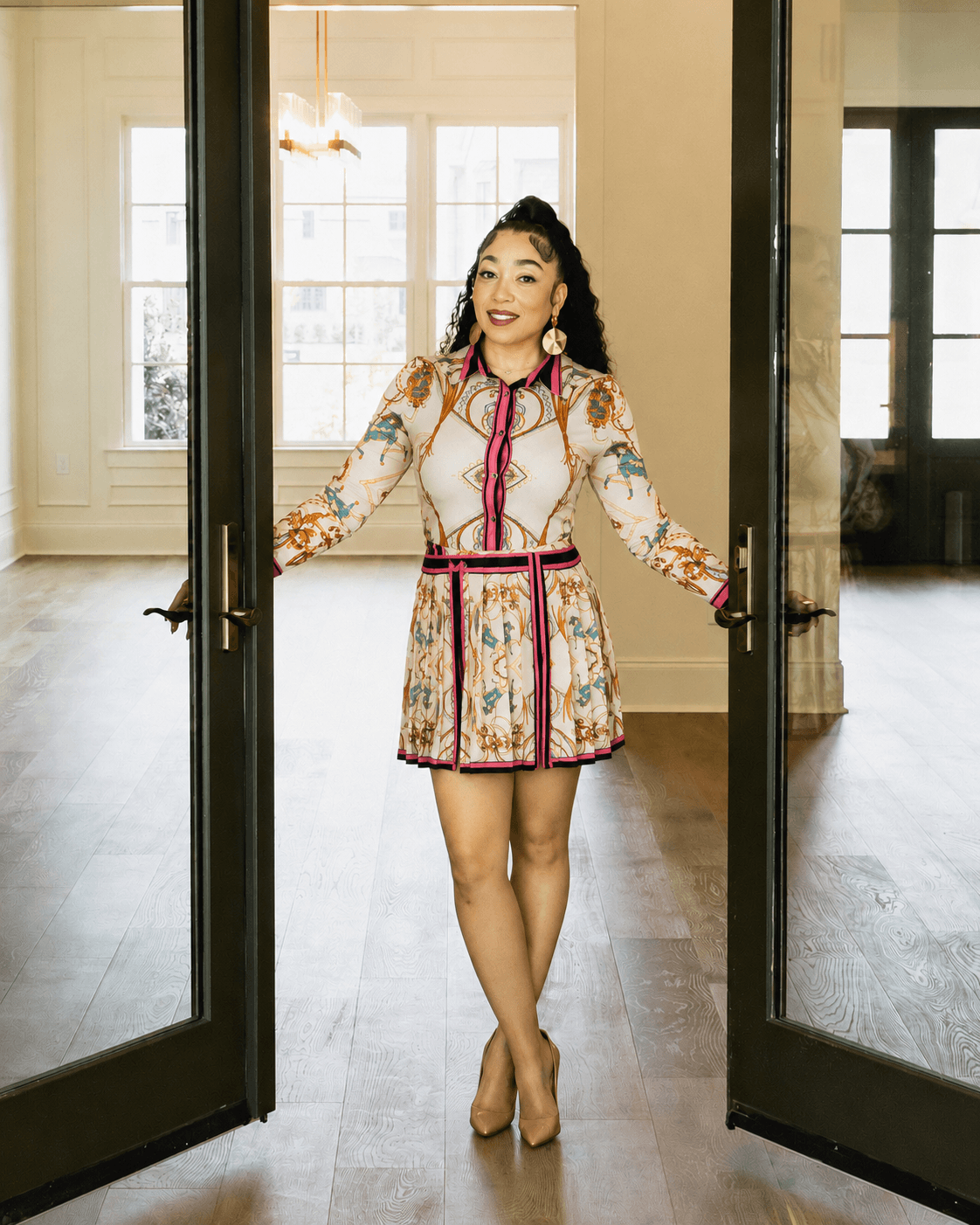 Woman in patterned dress standing in doorway, smiling, with large windows and wooden floor in background.
